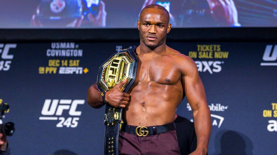 UFC 桌布 壁紙 — 一位肌肉發達的UFC拳手自信地站在稱重活動中，手持冠軍腰帶，背後有宣傳橫幅。