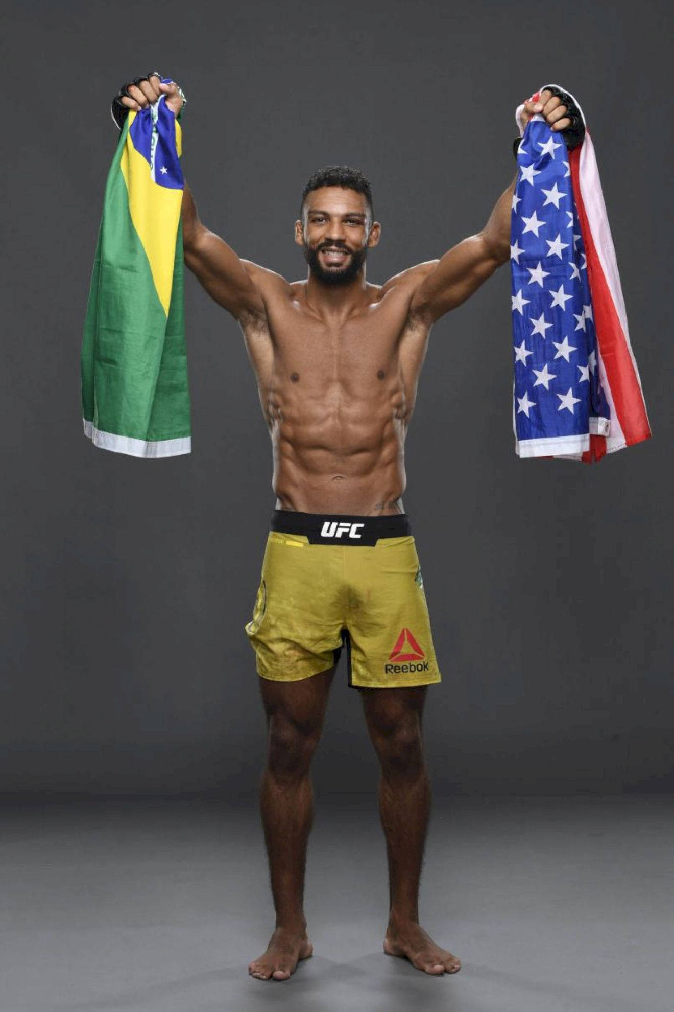 UFC 桌布 壁紙 — 一位肌肉發達的UFC拳手自豪地高舉巴西和美國國旗，在灰色攝影棚背景下露出勝利的笑容。