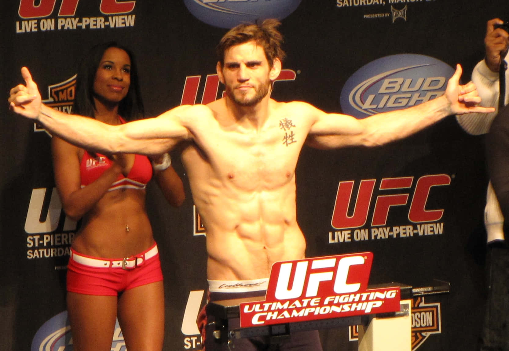 UFC 桌布 壁紙 — 一位肌肉發達的UFC拳擊手在品牌背景前，自信地在體重磅上擺姿勢，旁邊有一位微笑的擂台女郎。