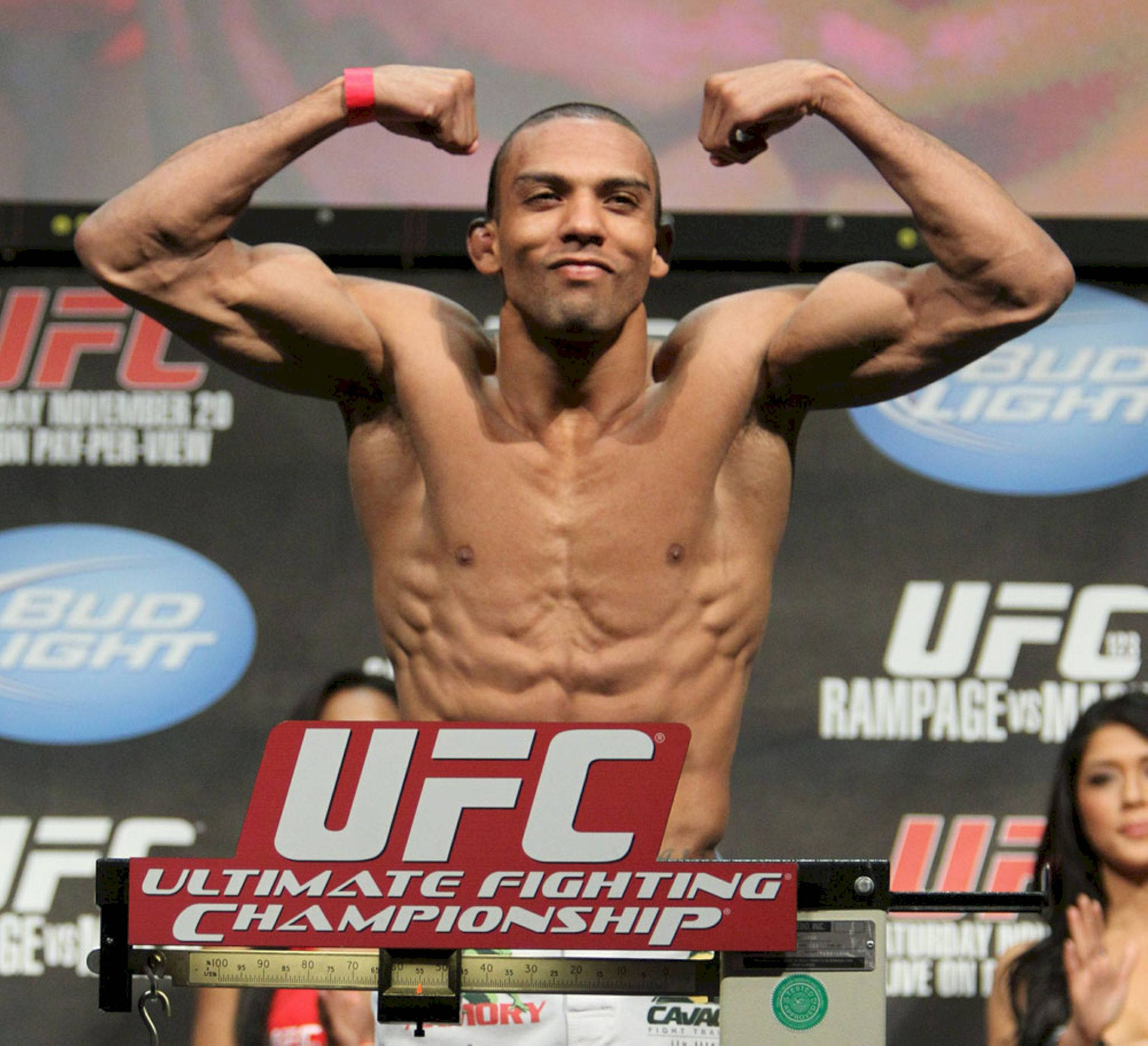 MMA 桌布 壁紙 — 一位肌肉發達的UFC拳擊手在稱重活動中，在帶有品牌橫幅的背景前，在秤上彎曲手臂。