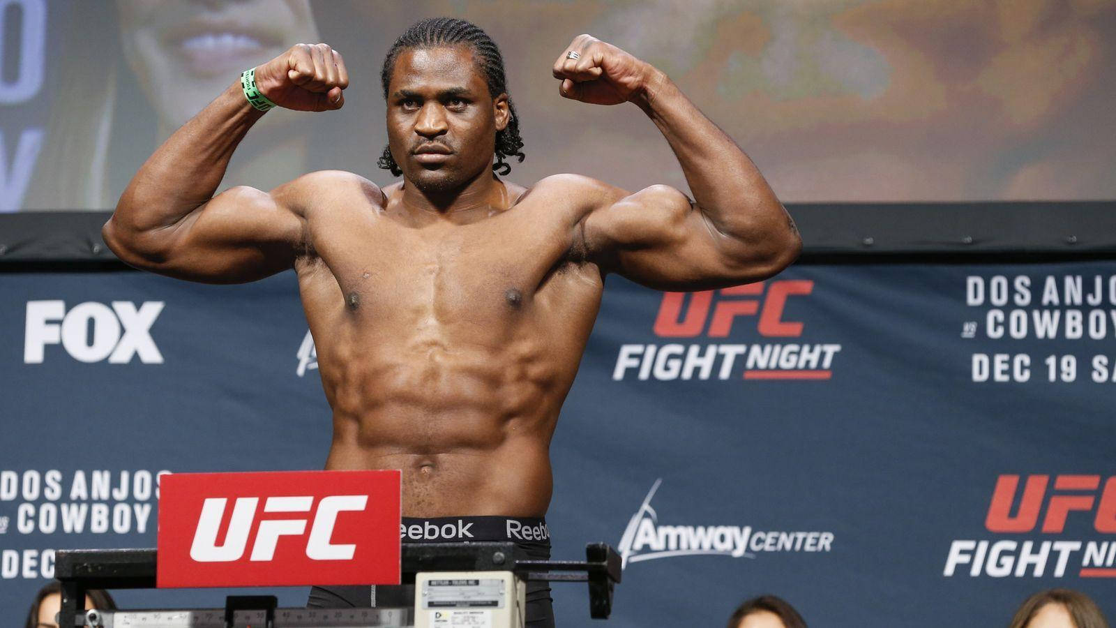 MMA 桌布 壁紙 — 一位肌肉發達的UFC拳擊手在稱重活動中，在體重計上展示他的二頭肌，UFC品牌和贊助商標誌清晰可見。