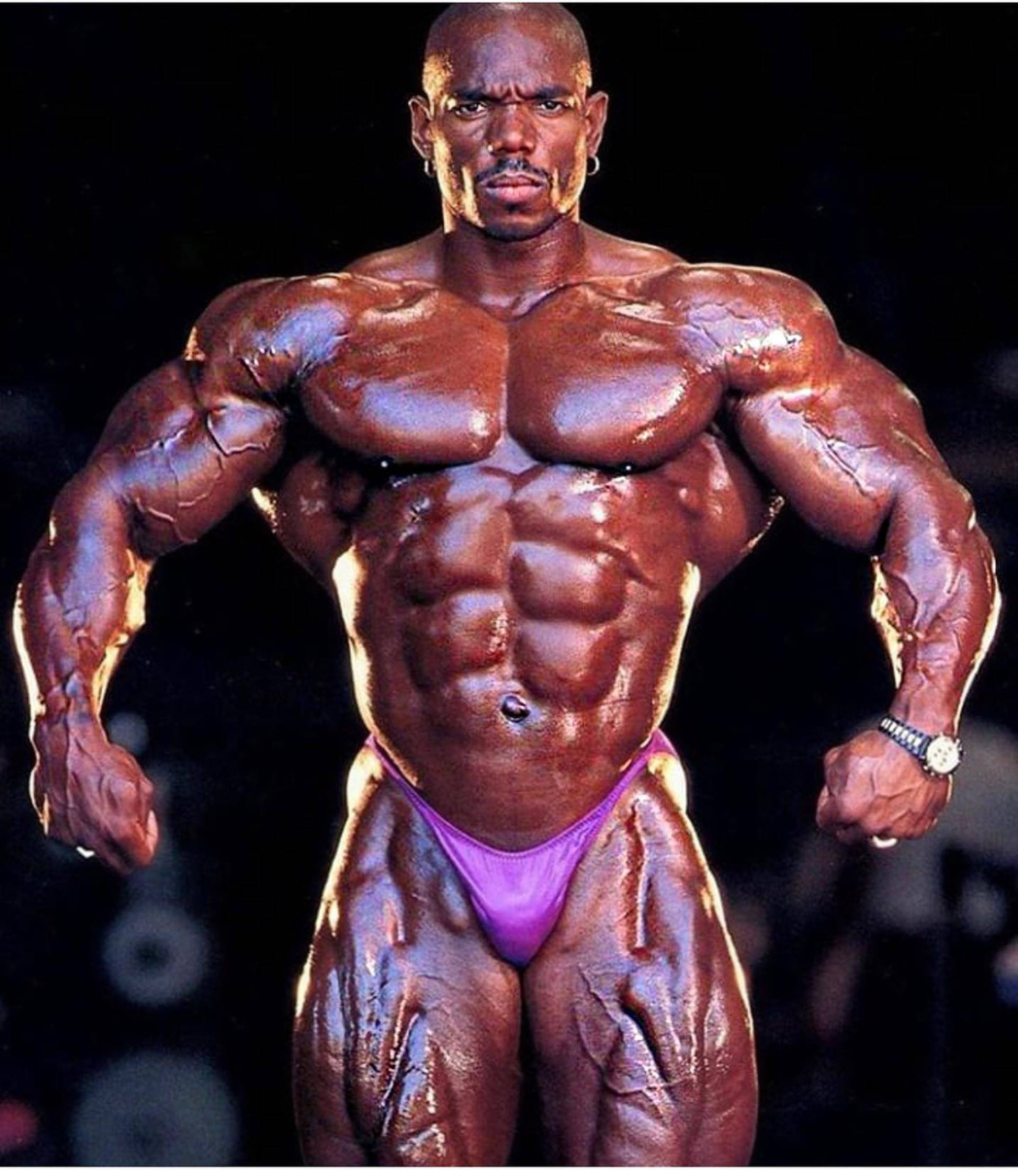 flex wheeler 桌布 壁紙 — 一位肌肉發達的健美運動員，皮膚塗油，在深色背景下穿著紫色比賽褲，擺出強烈的姿勢。