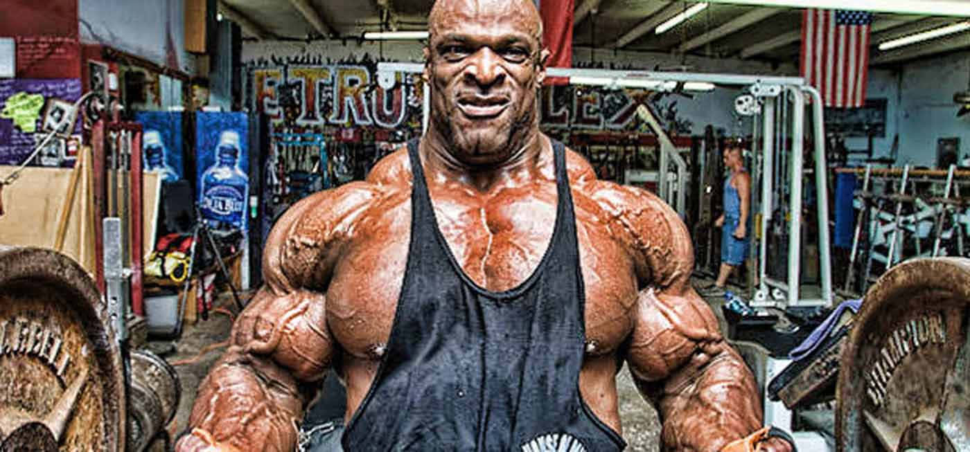 運動 桌布 壁紙 — 一位肌肉發達的健美運動員，皮膚閃閃發光，在雜亂的健身房中自信地擺姿勢，周圍是啞鈴和器材。