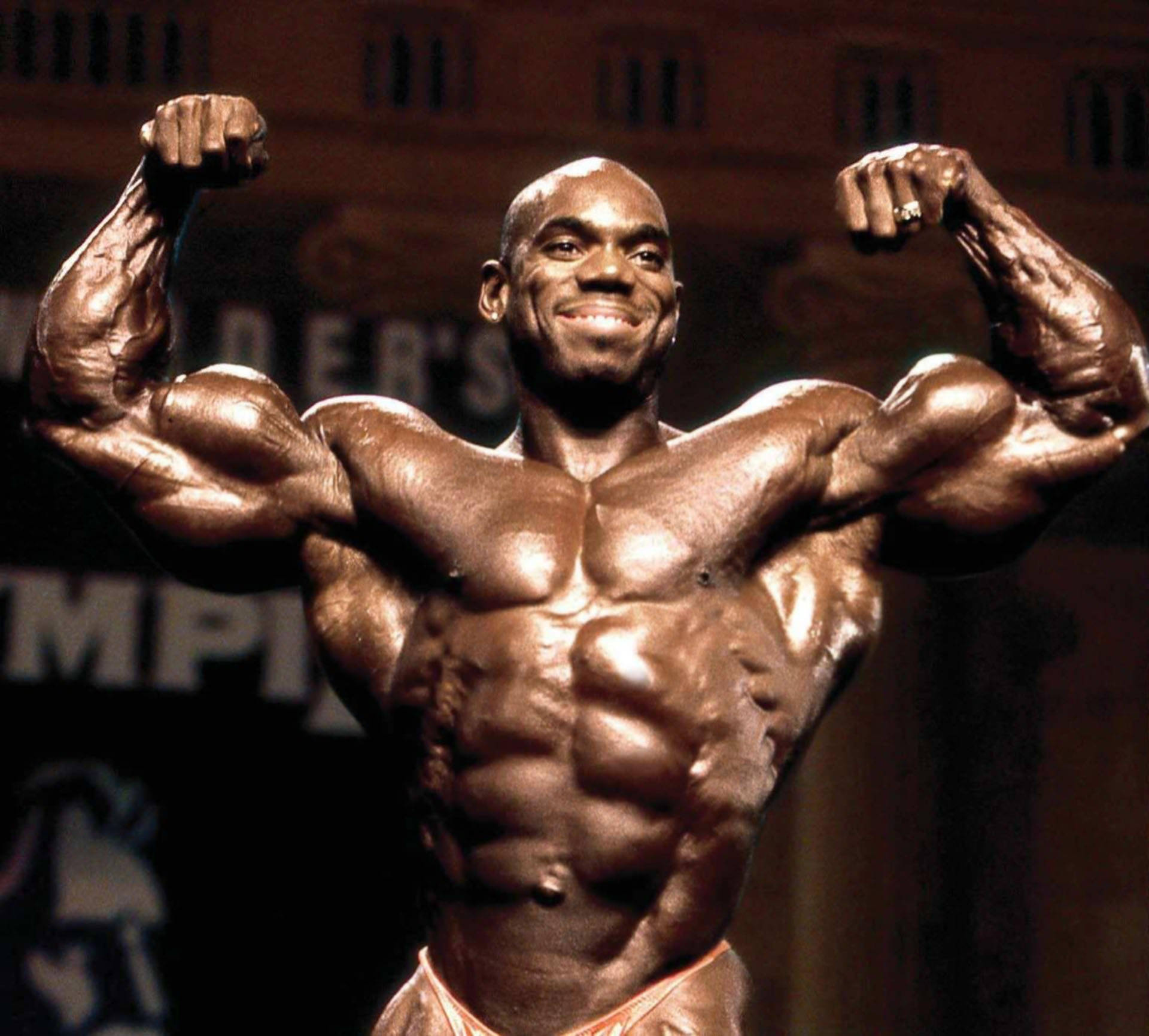 flex wheeler 桌布 壁紙 — 一位肌肉發達的健美運動員在舞台燈光下自信微笑，繃緊手臂和胸部。