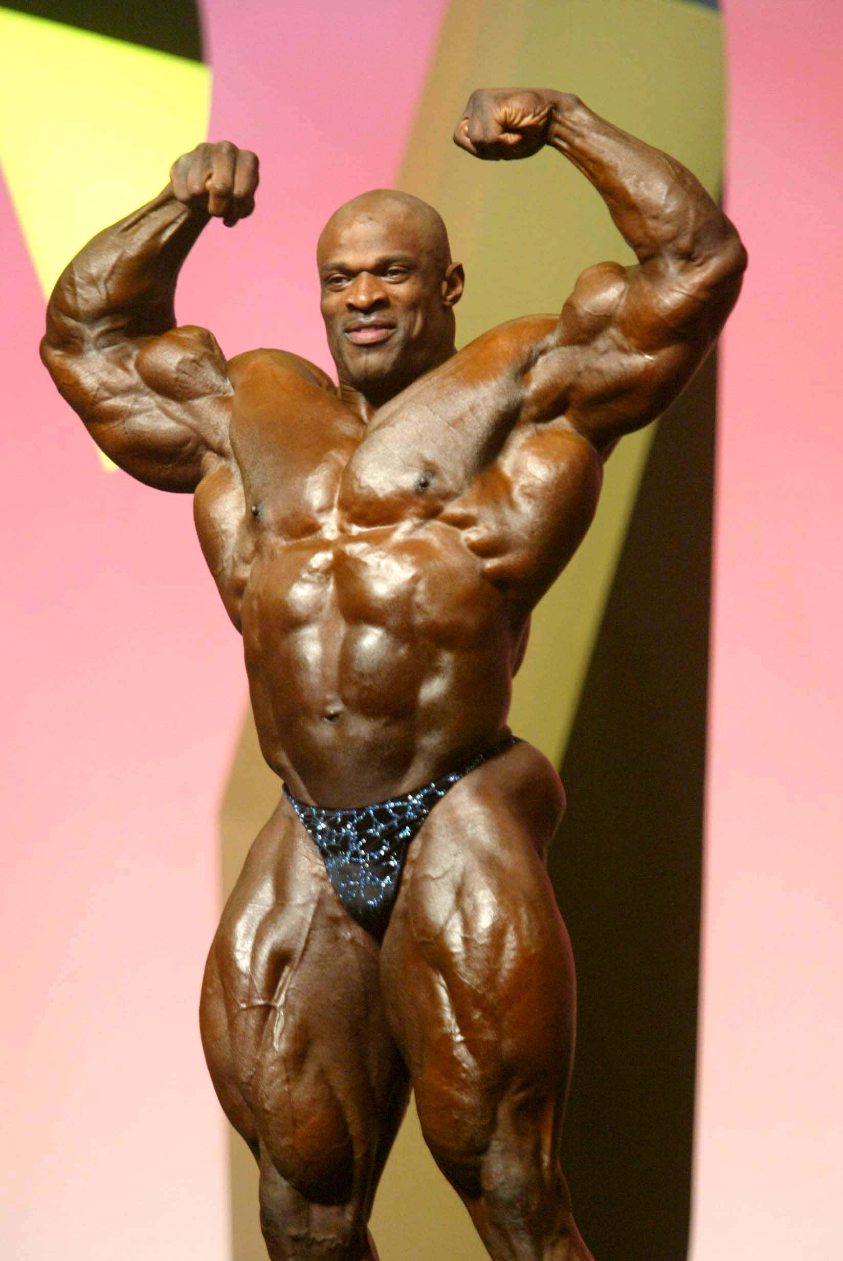 運動 桌布 壁紙 — 一位肌肉發達的健美運動員在色彩繽紛的舞台上，於明亮燈光下彎曲手臂。