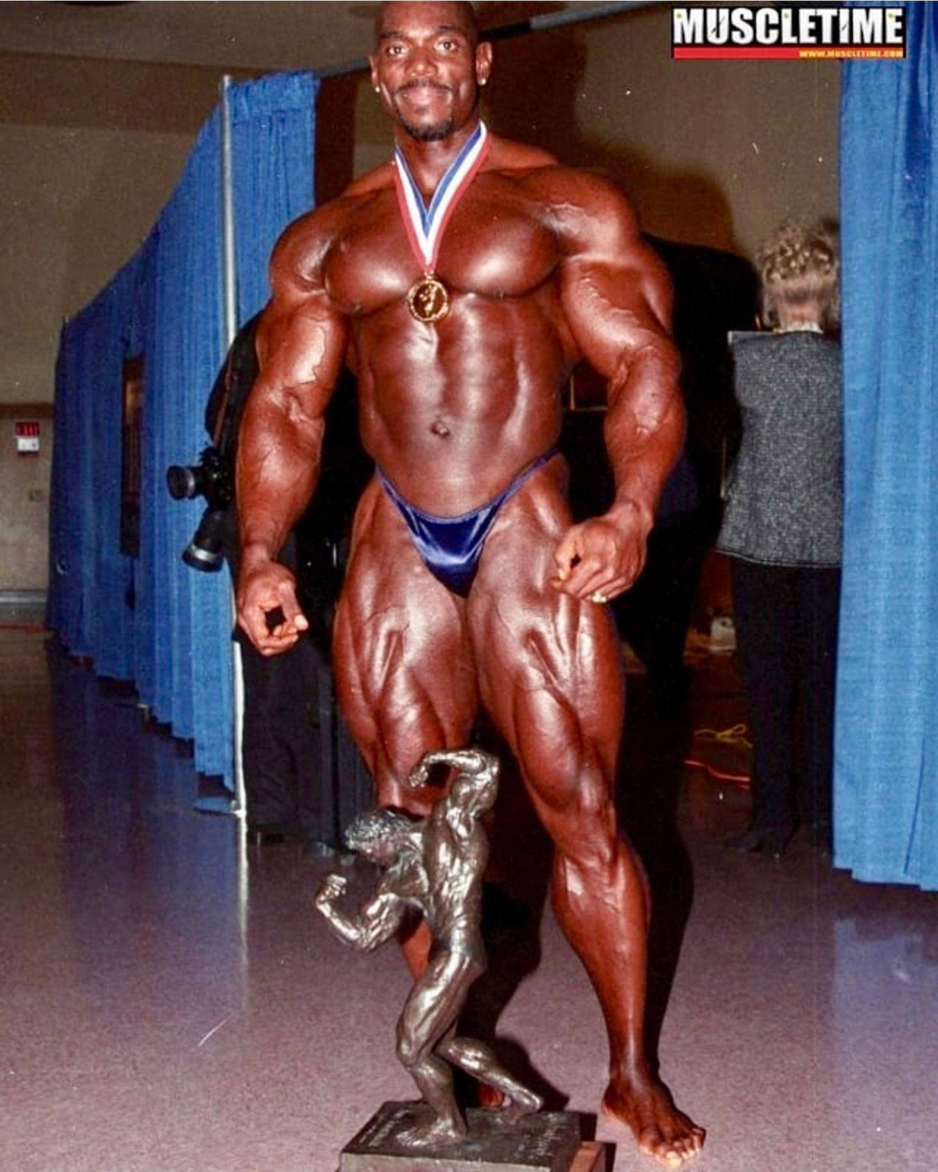 flex wheeler 桌布 壁紙 — 一位肌肉發達的健美運動員在藍色窗簾的比賽場館中自豪地展示金牌和獎盃。