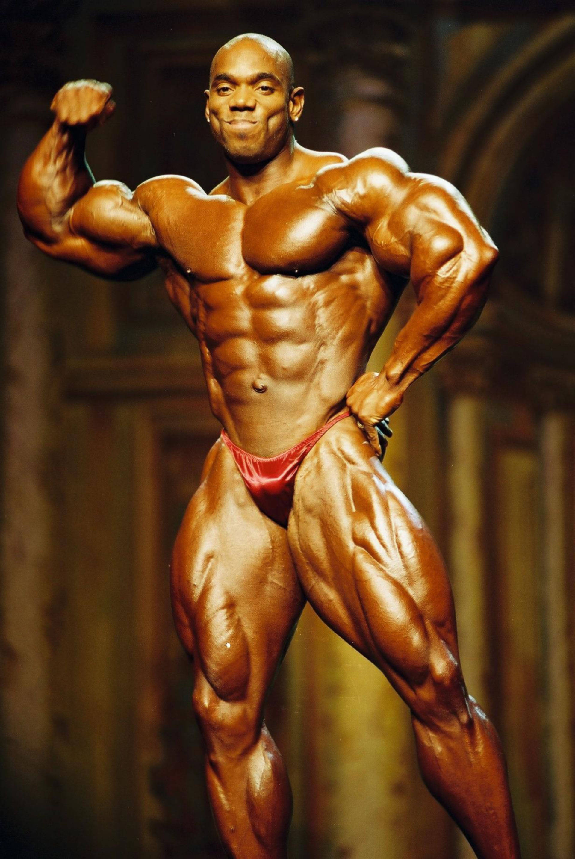 flex wheeler 桌布 壁紙 — 一位肌肉發達的健美運動員穿著紅色比賽褲，在舞台燈光下展示他線條分明的體格。