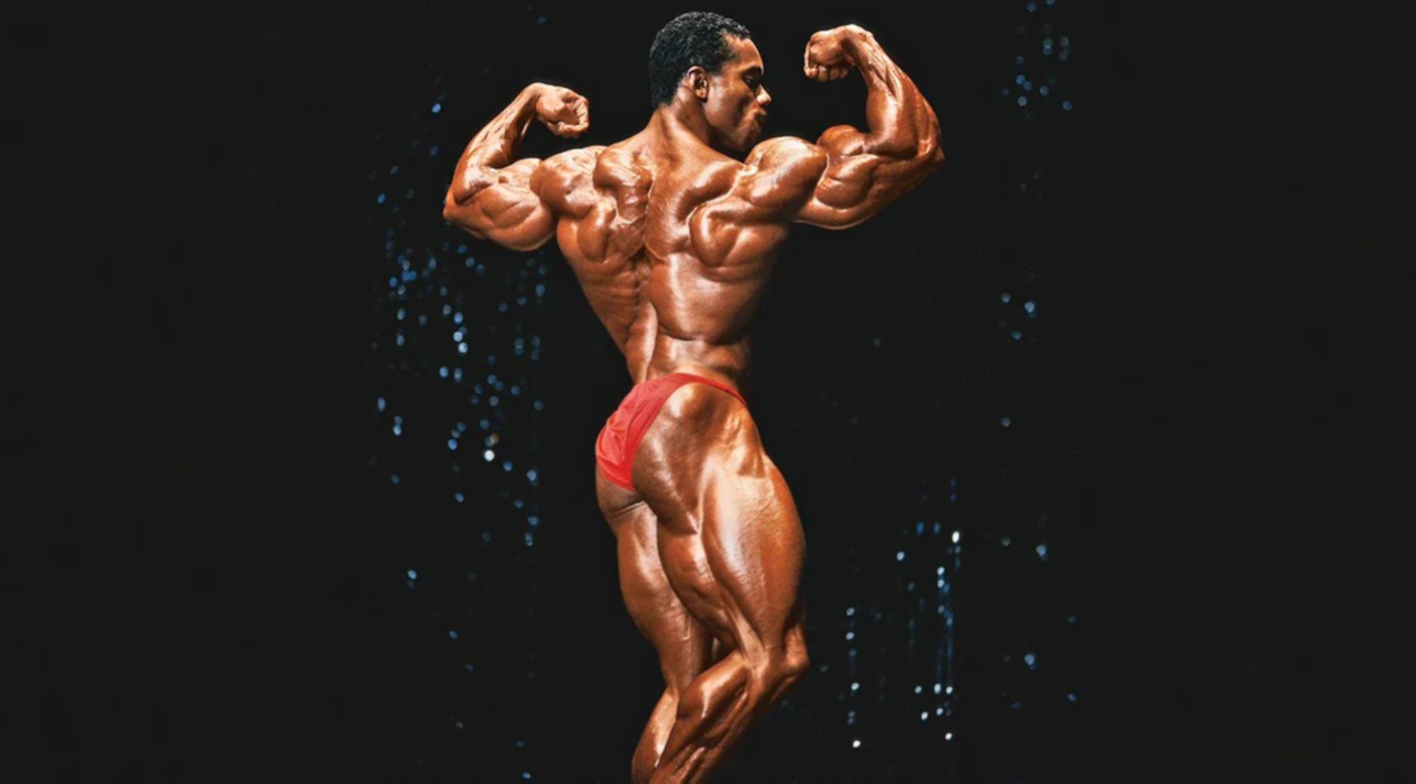弗萊克斯·惠勒 桌布 壁紙 — 一位肌肉發達的健美運動員穿著紅色比賽褲，在帶有閃爍燈光的黑暗舞台上擺姿勢。