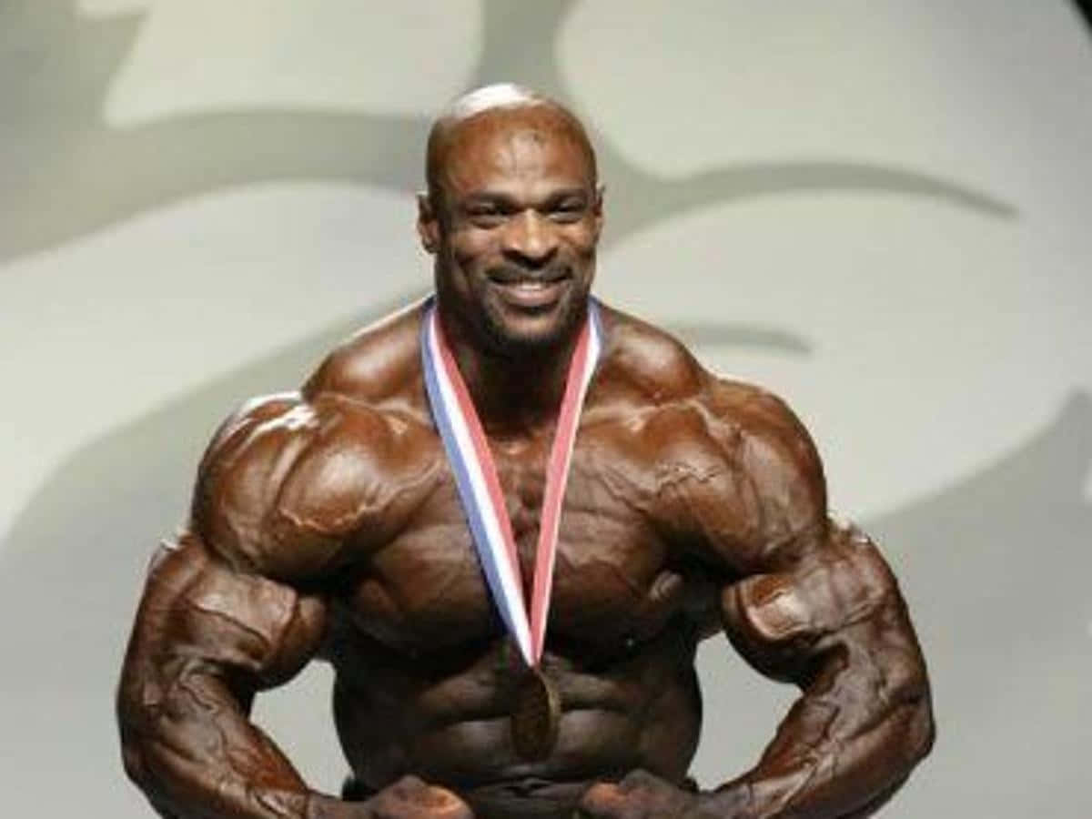 運動 桌布 壁紙 — 一位肌肉發達的健美運動員脖子上掛著金牌，在舞台上自信地擺姿勢，肌肉線條分明，露出自豪的笑容。