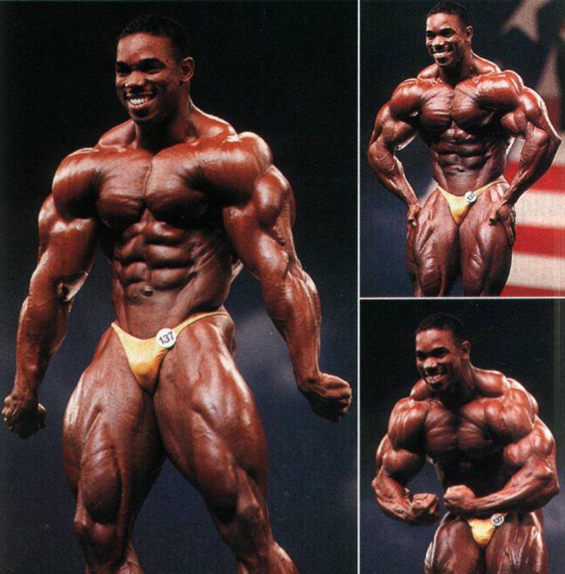 flex wheeler 桌布 壁紙 — 一位肌肉發達的健美運動員自信地穿著黃色比賽褲，在黑暗背景前擺姿勢，背景有美國國旗。