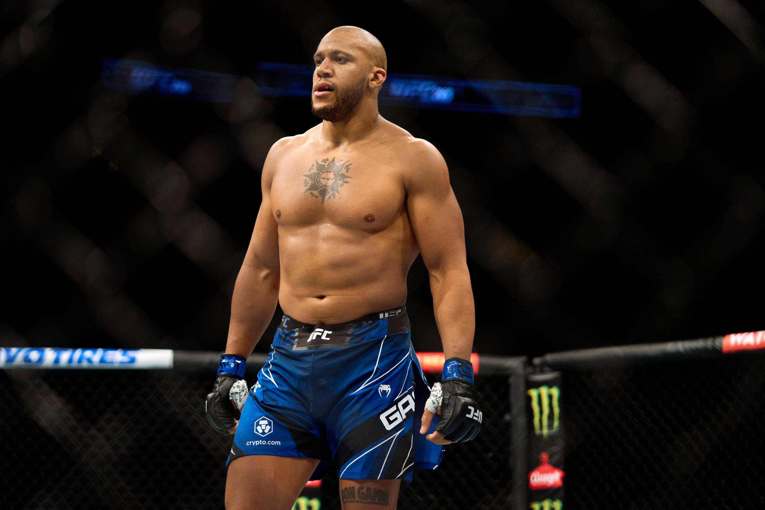 MMA 桌布 壁紙 — 一位肌肉發達的光頭戰士，胸口有刺青，站在燈光昏暗的MMA八角籠內，穿著藍色短褲和手套。