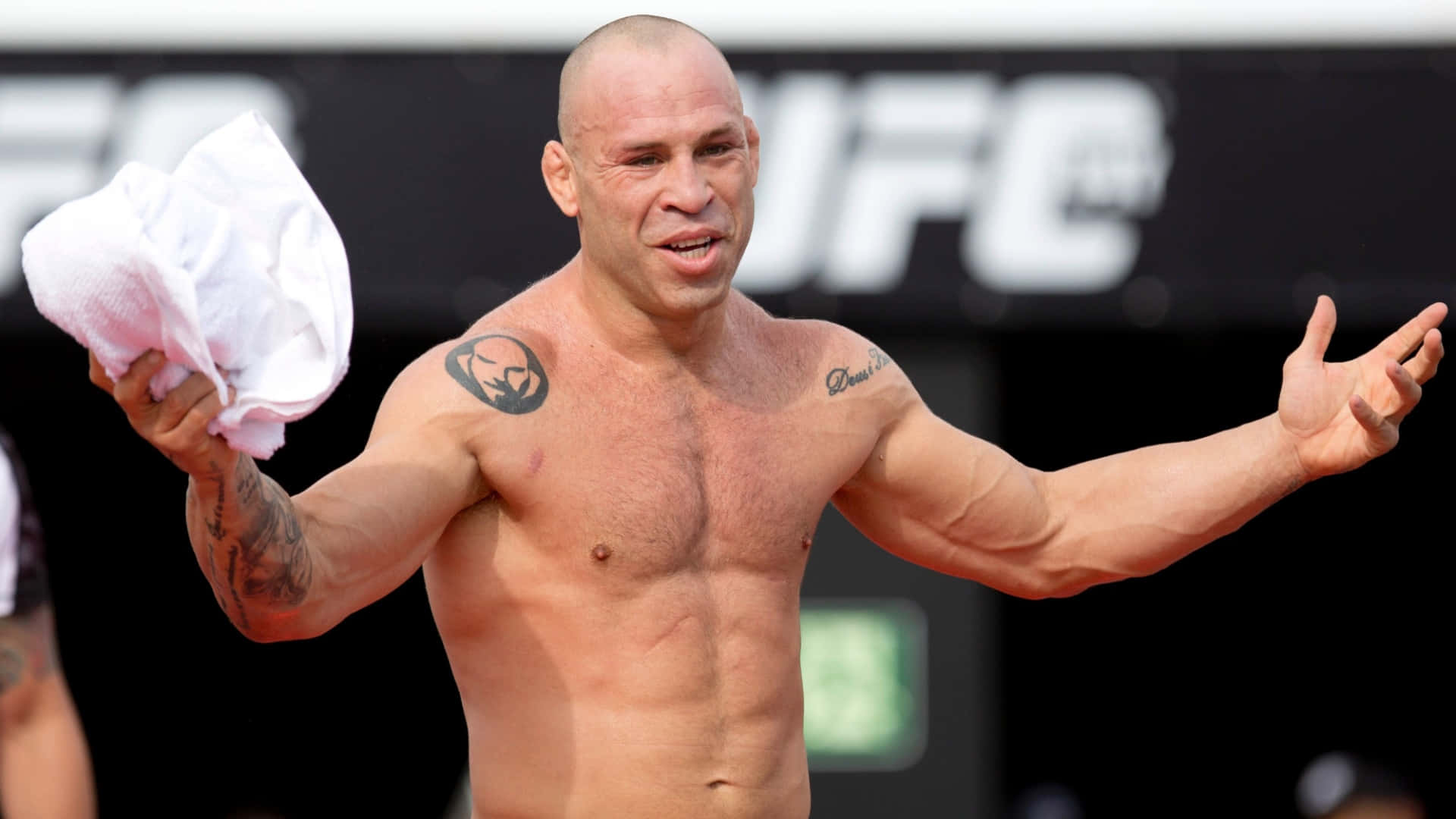 UFC 桌布 壁紙 — 一位肌肉發達的光頭戰士帶有刺青，在模糊的UFC背景下戲劇性地做手勢，同時拿著毛巾。