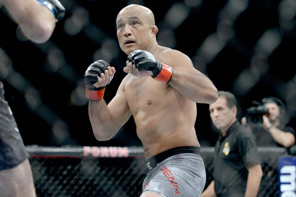 UFC 桌布 壁紙 — 一位肌肉發達的光頭戰士穿著灰色短褲和黑色手套，在昏暗的八角籠內揮拳，圍欄上可見「FORUM」字樣。