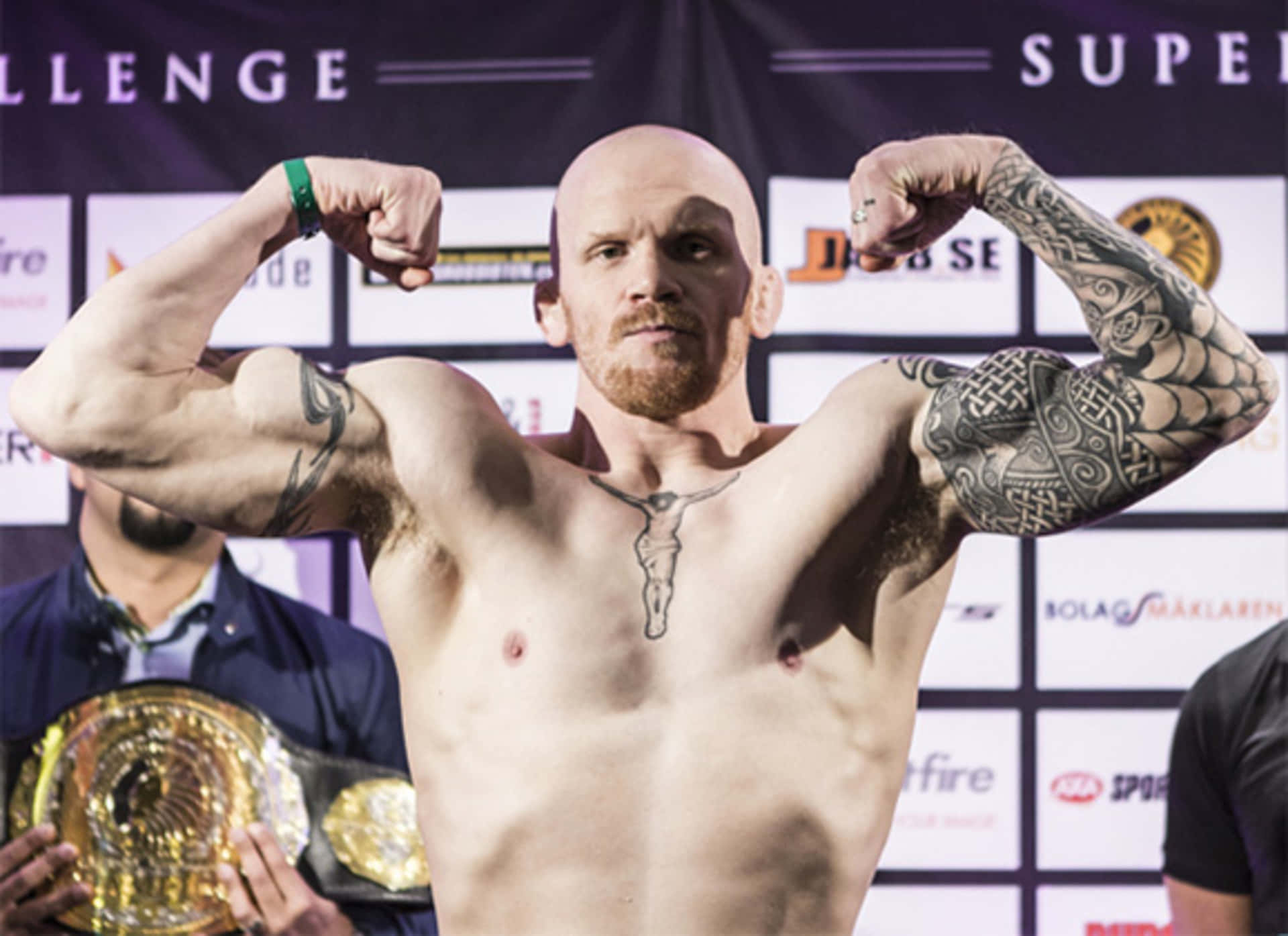 MMA 桌布 壁紙 — 一位肌肉發達的光頭拳手，身上有刺青，在稱重活動中戴著冠軍腰帶，彎曲手臂。