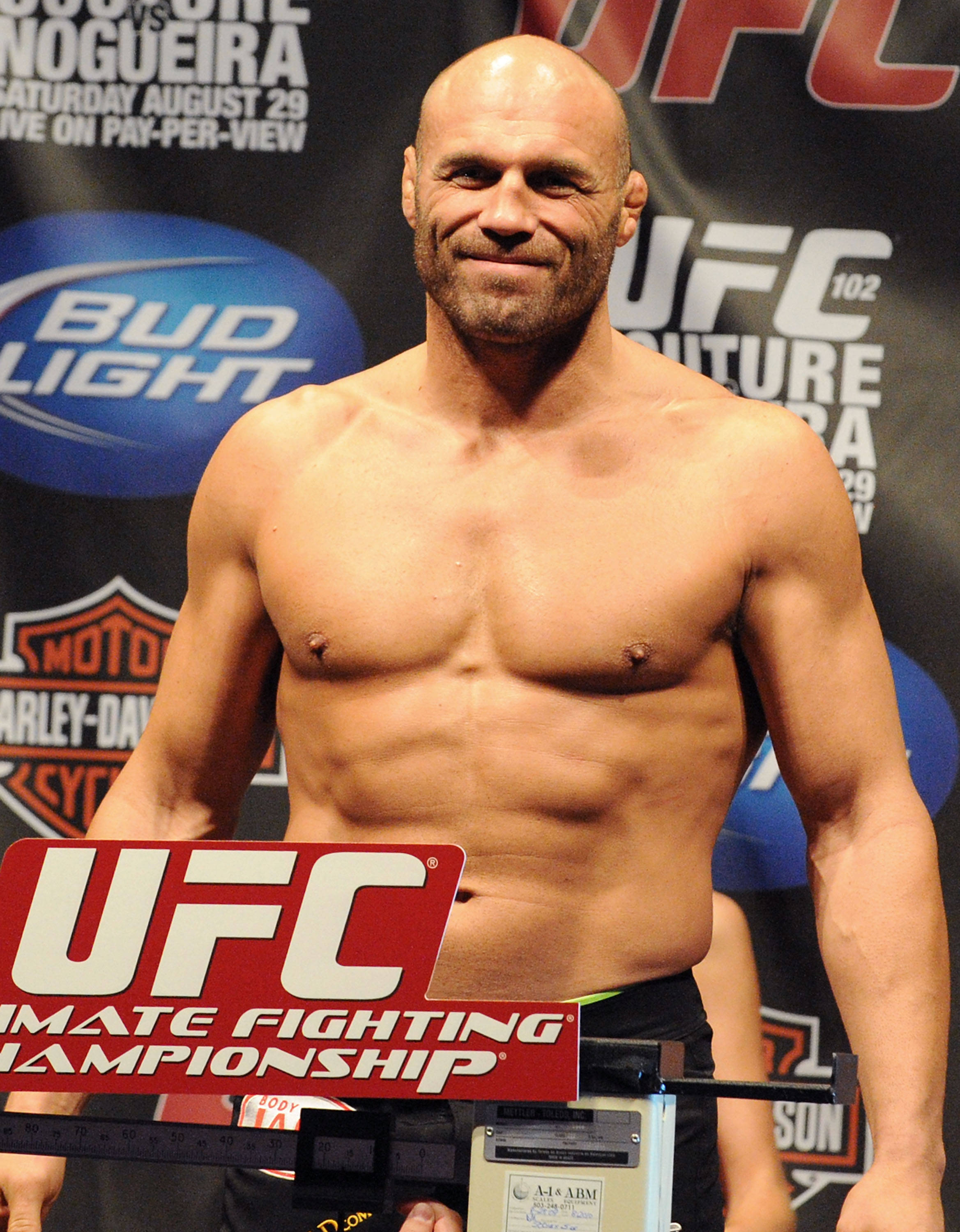 MMA 桌布 壁紙 — 一位肌肉發達的光頭拳手在 UFC 稱重活動中站在體重磅上，自信地對著鏡頭微笑。
