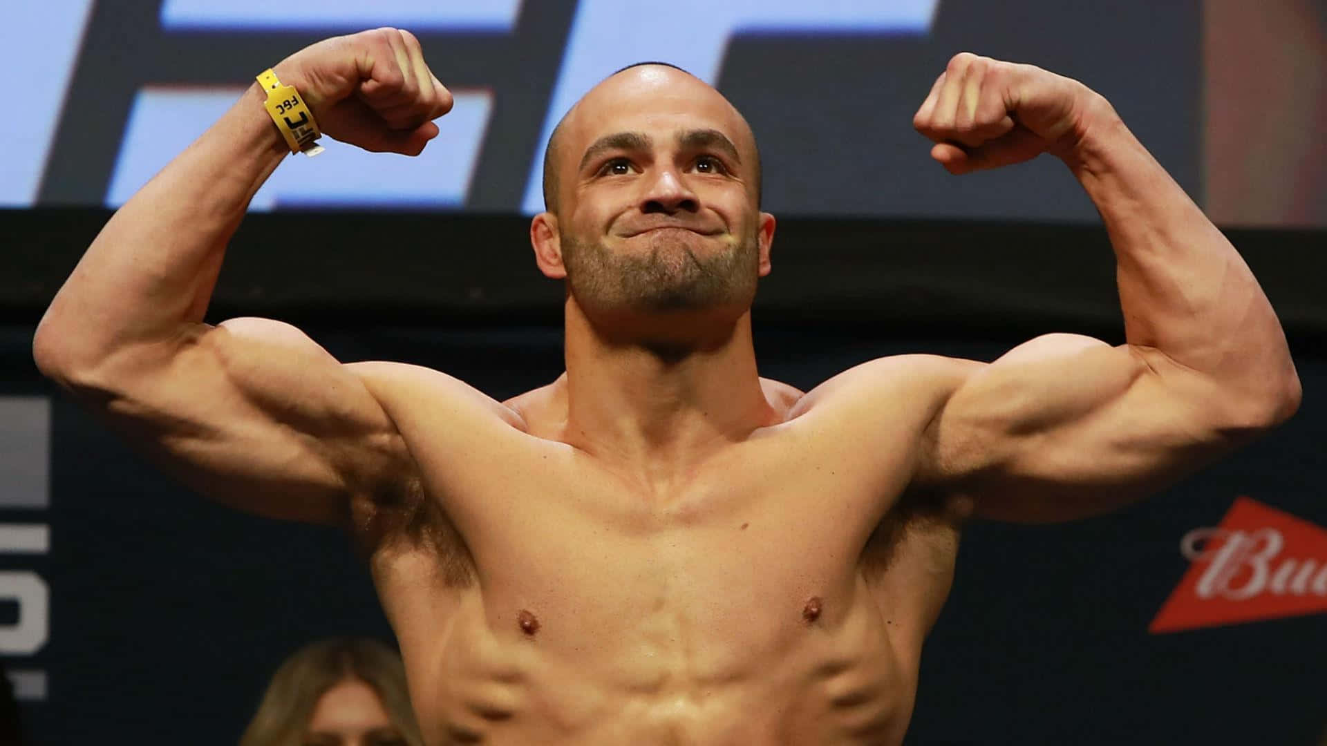 UFC 桌布 壁紙 — 一位肌肉發達的光頭拳手在明亮的場館燈光下，於稱重活動中自信地微笑並彎曲雙臂。