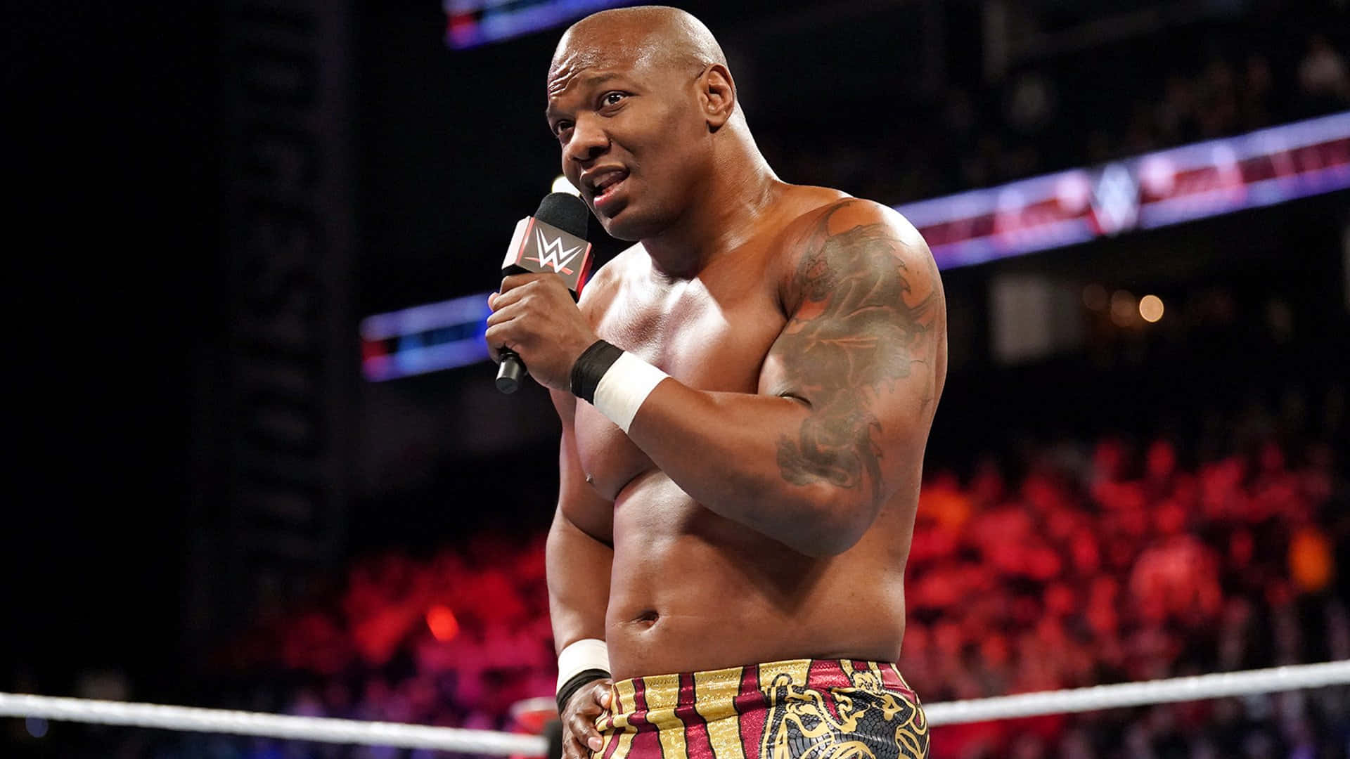 WWE 桌布 壁紙 — 一位肌肉發達的光頭摔角手帶著刺青，在擂台上拿著WWE麥克風，身後紅光照耀的觀眾為他歡呼。