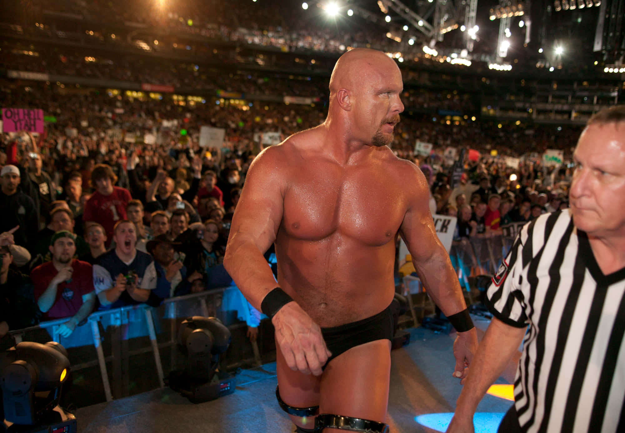 WWE 桌布 壁紙 — 一位肌肉發達的光頭摔角手穿著黑色三角褲，自信地與裁判一起走過一個燈火通明、充滿咆哮粉絲的競技場。