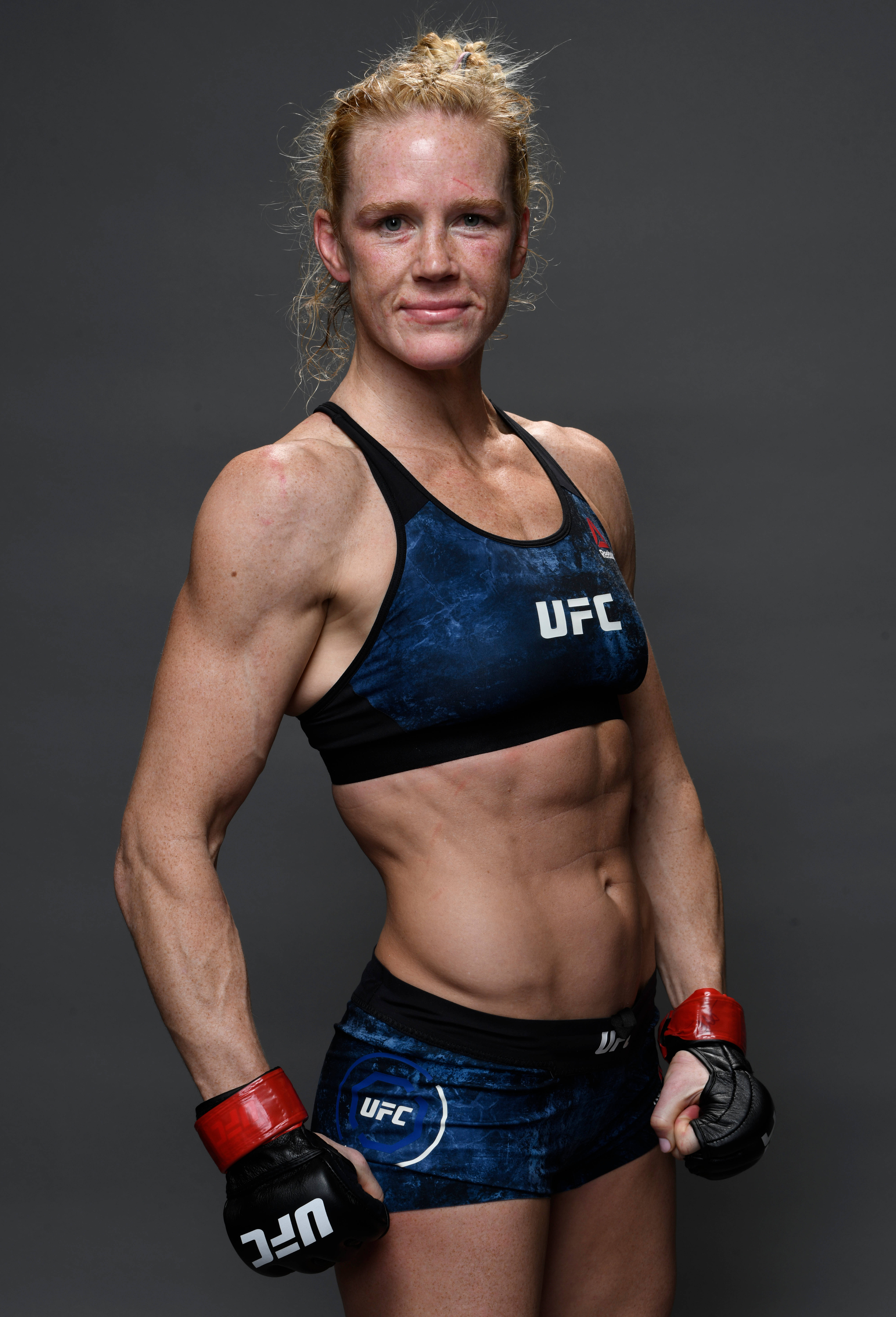 人像動態 桌布 壁紙 — 一位肌肉發達的女性UFC拳手，身穿藍色裝備和紅色手套，在深灰色工作室背景前自信地擺姿勢。