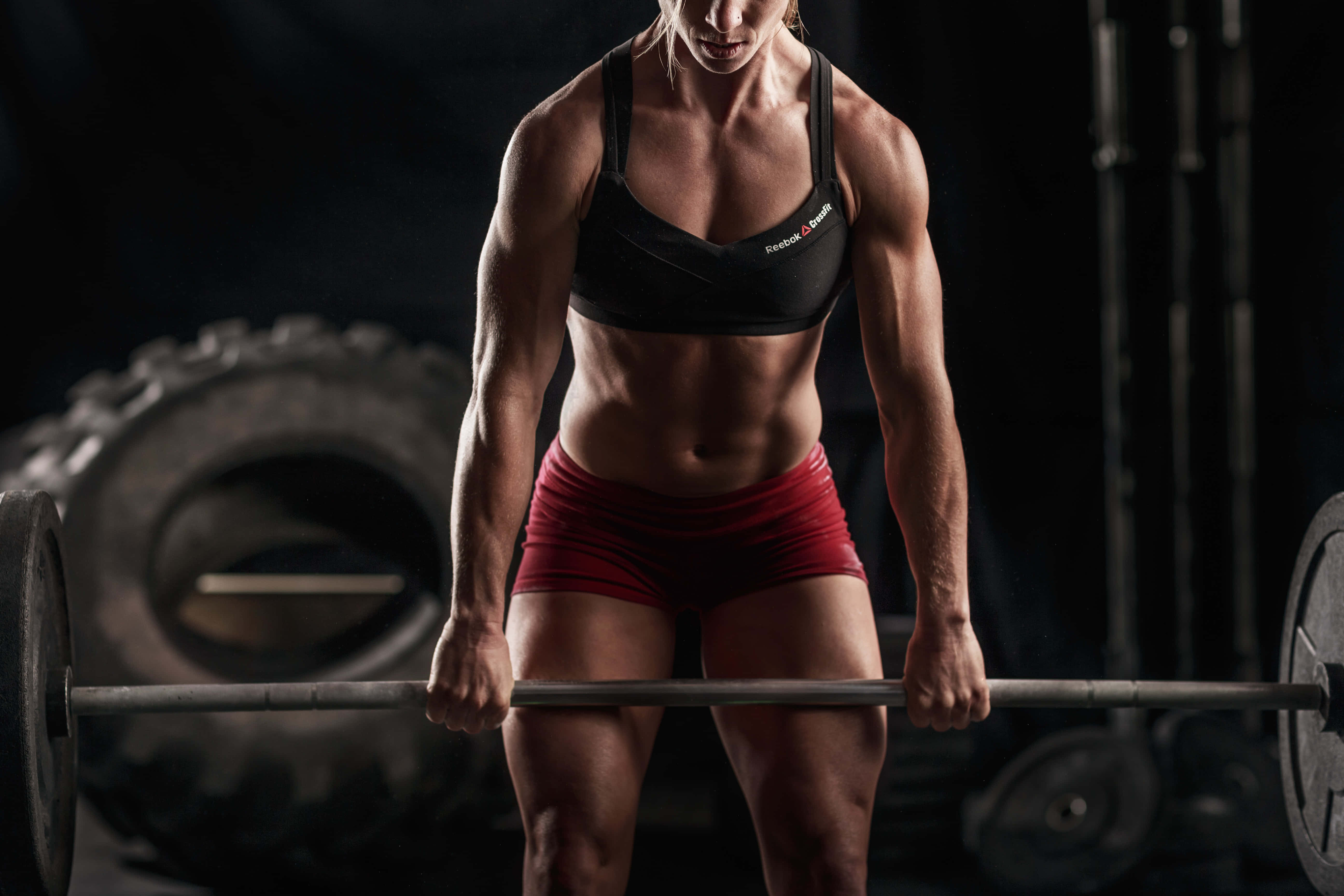 Barbell 桌布 壁紙 — 一位肌肉發達的女性在健身房緊握槓鈴，在戲劇性的燈光下準備舉重，神情專注。