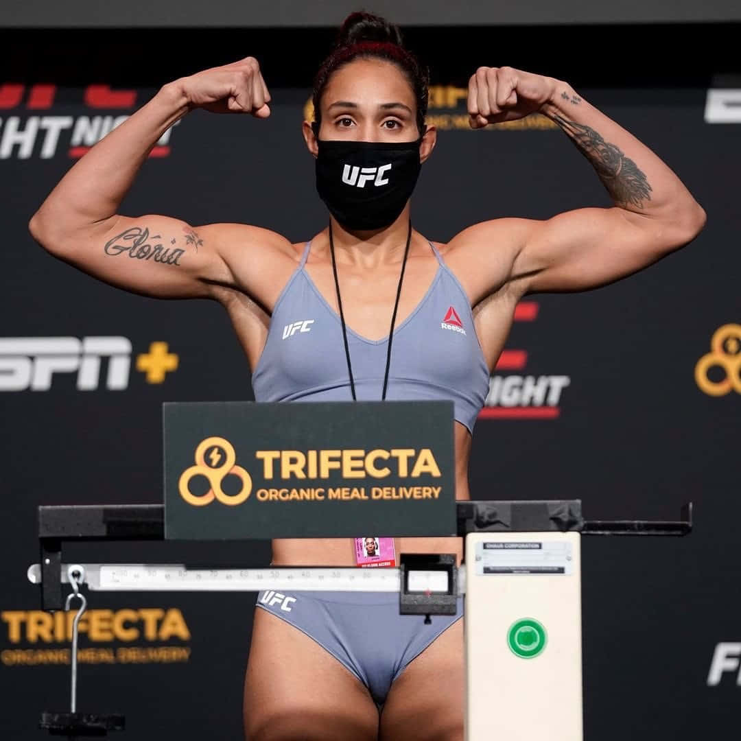 MMA 桌布 壁紙 — 一位肌肉發達的女拳手在UFC稱重秤上彎曲她的二頭肌，戴著黑色口罩和灰色運動裝備。