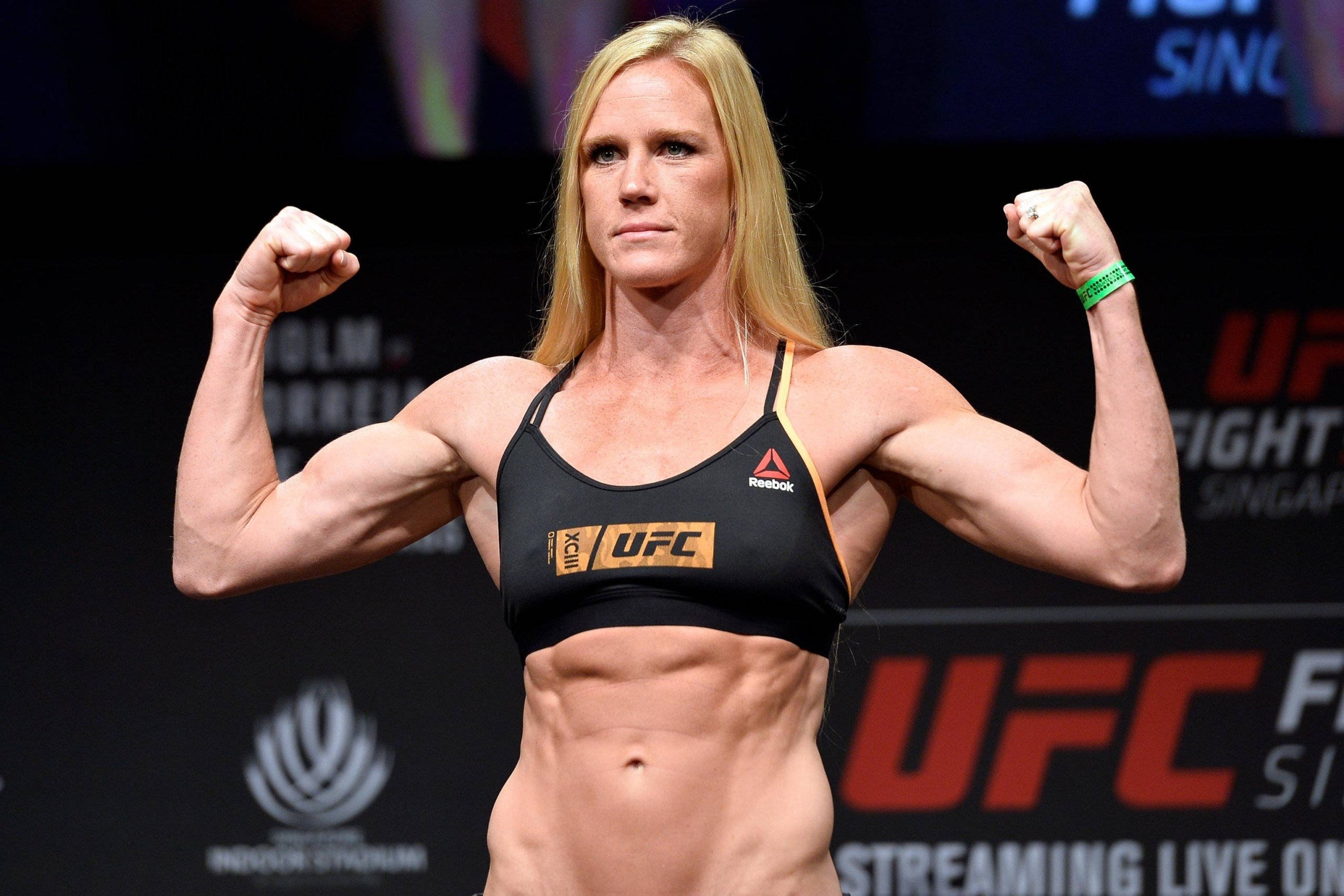 MMA 桌布 壁紙 — 一位肌肉發達的女拳手在明亮的競技場燈光下，穿著黑色UFC運動內衣彎曲手臂。