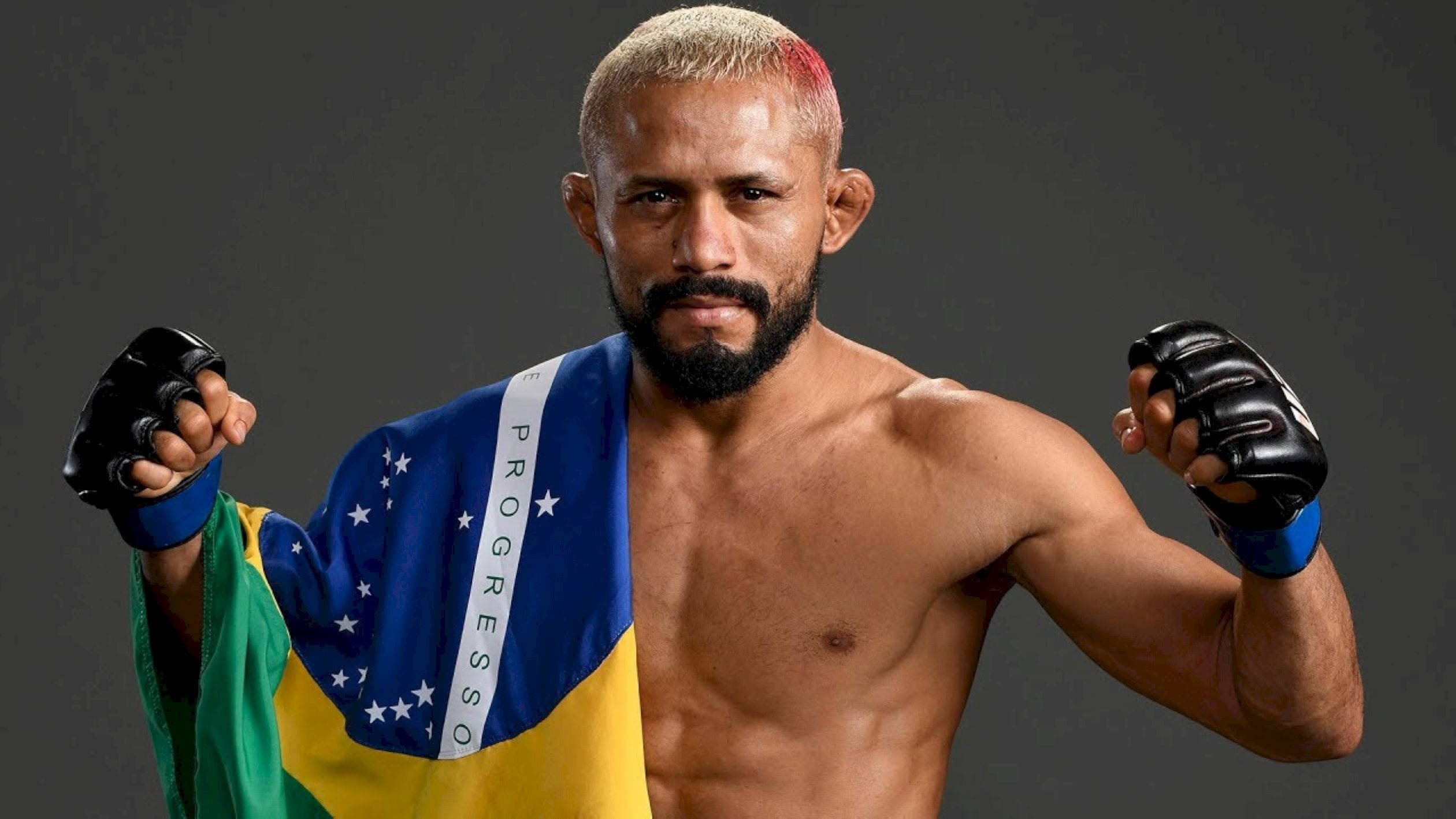UFC 桌布 壁紙 — 一位肌肉發達的巴西戰士，頭髮上有一條紅色條紋，自信地站著，戴著黑色手套，披著巴西國旗。