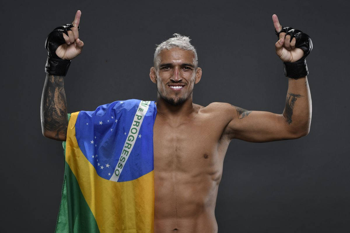 UFC 桌布 壁紙 — 一位肌肉發達的巴西戰士在黑色攝影棚背景前，披著巴西國旗，自豪地微笑並舉起拳頭。