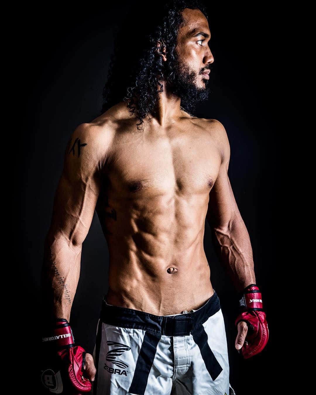 Fight 桌布 壁紙 — 一位肌肉發達的戰士，留著長捲髮，戴著紅色手套，側身站在黑色背景前，展示出清晰的腹肌和刺青。