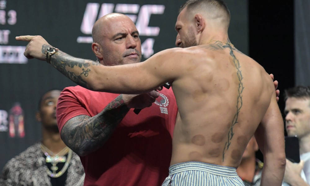 UFC 桌布 壁紙 — 一位肌肉發達的戰士，背上有刺青和拔罐印記，站在一位身穿紅衫的光頭男子面前，該男子在UFC賽事中向前指點。