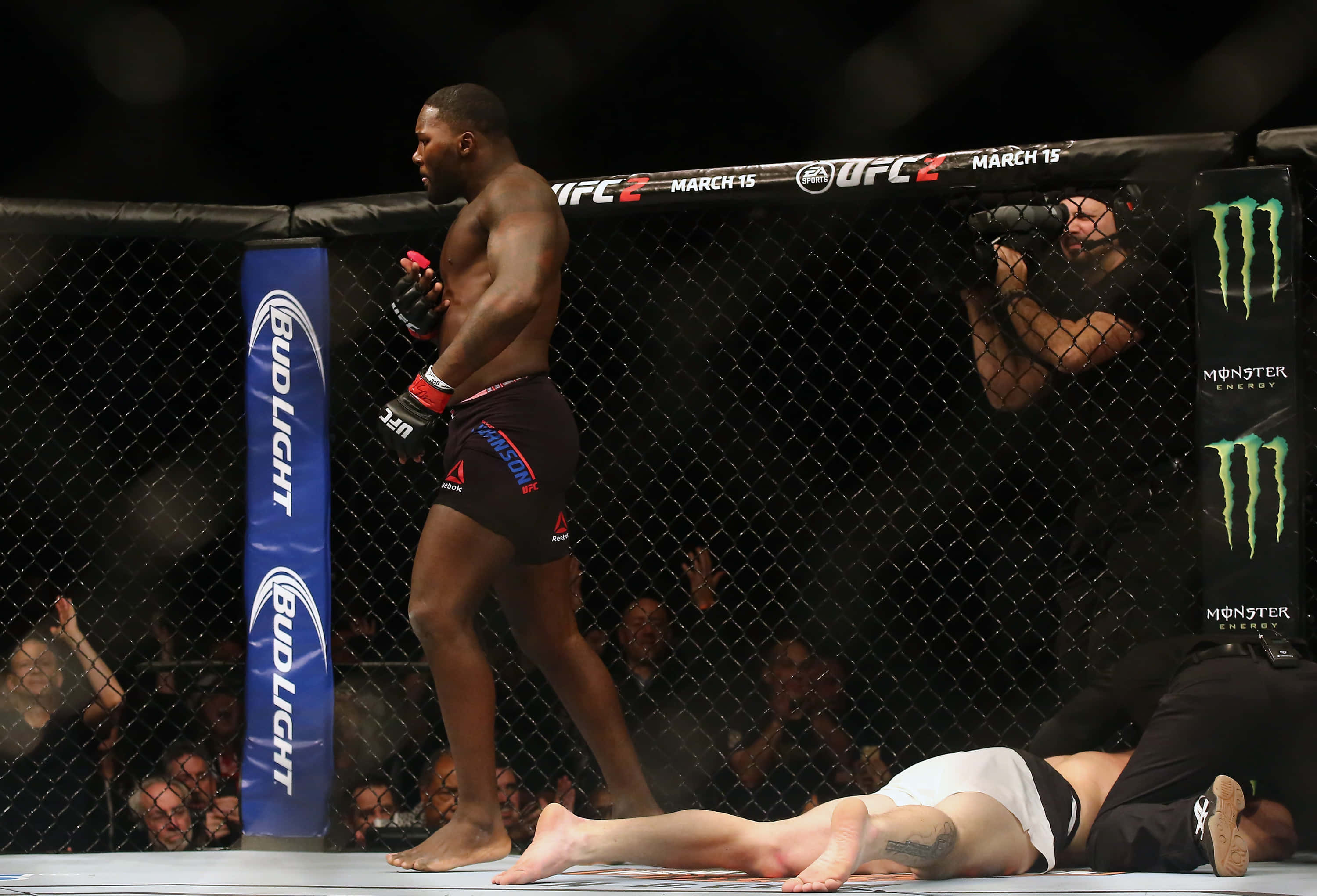 MMA 桌布 壁紙 — 一位肌肉發達的戰士在UFC八角籠中勝利站立，而他的對手在明亮的場館燈光下躺在墊子上一動不動。