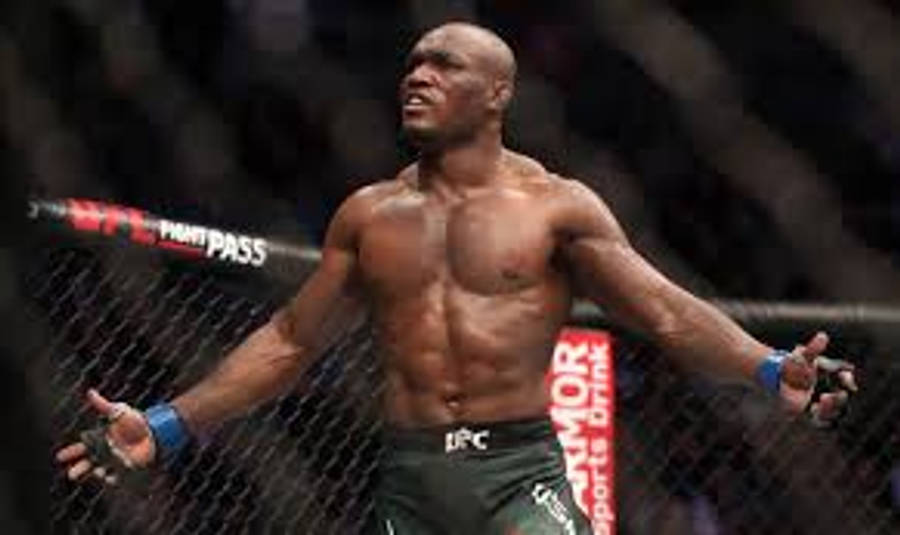 UFC 桌布 壁紙 — 一位肌肉發達的戰士在明亮的場館燈光下，於UFC八角籠中張開雙臂，勝利地站立。