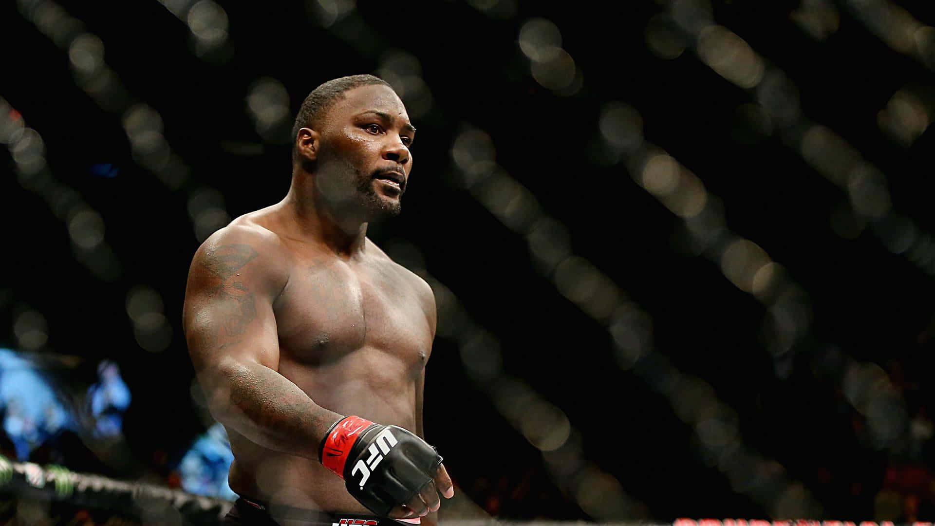 MMA 桌布 壁紙 — 一位肌肉發達的戰士在昏暗的八角籠中專注站立，背景是模糊的籠網圍欄。
