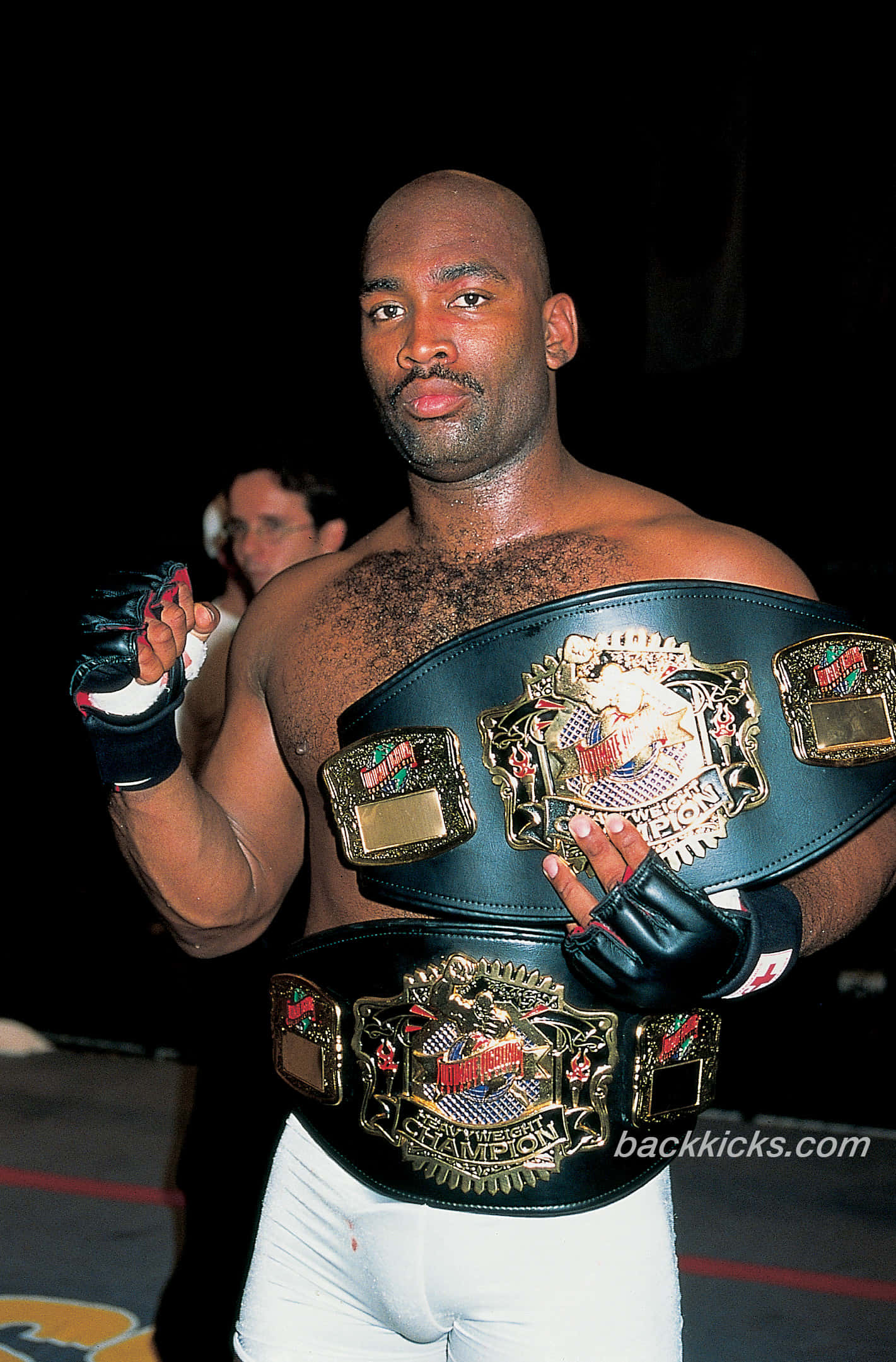 MMA 桌布 壁紙 — 一位肌肉發達的戰士在深色背景下自豪地展示兩條冠軍腰帶，表情專注。