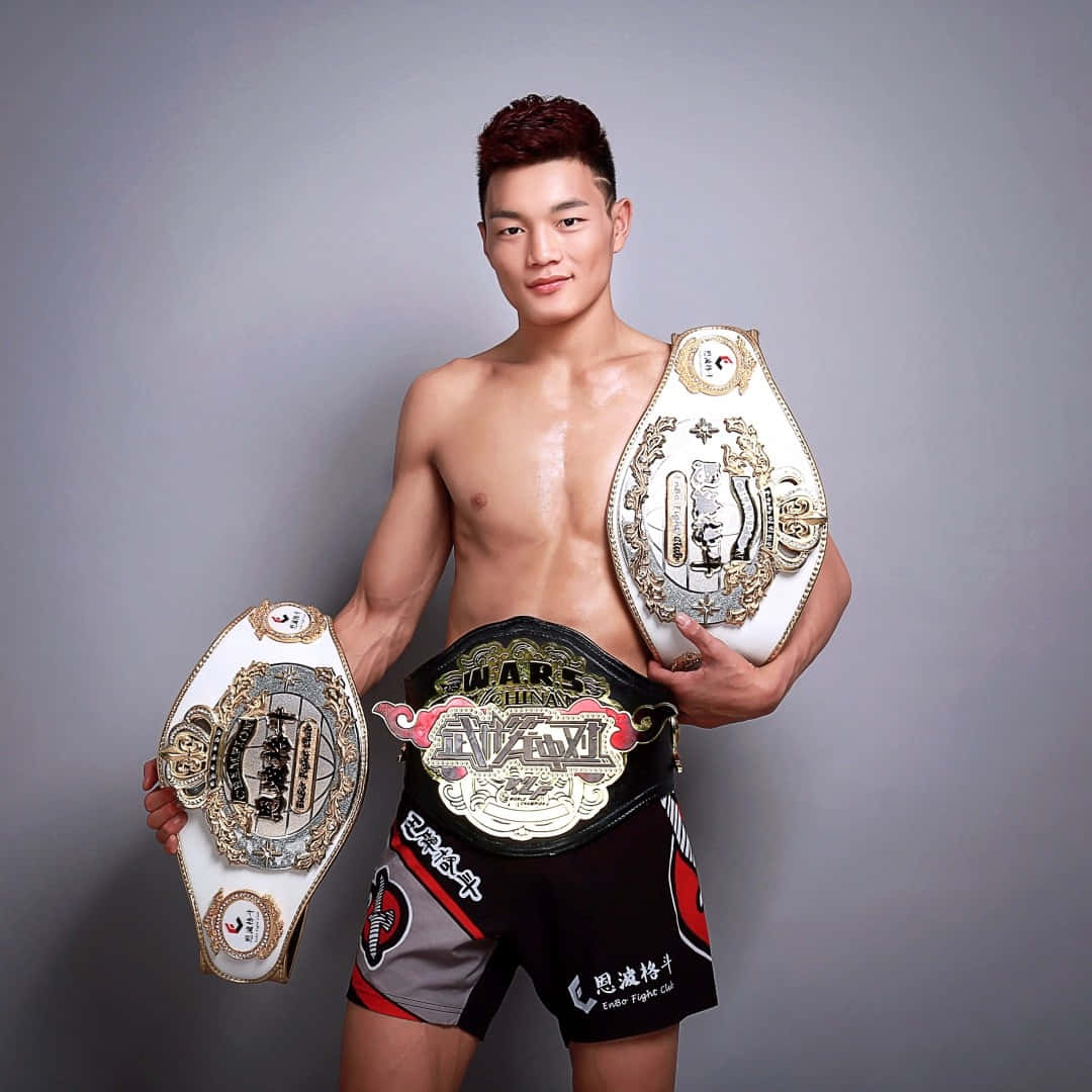 MMA 桌布 壁紙 — 一位肌肉發達的戰士在純灰色工作室背景下自豪地展示三條華麗的冠軍腰帶。