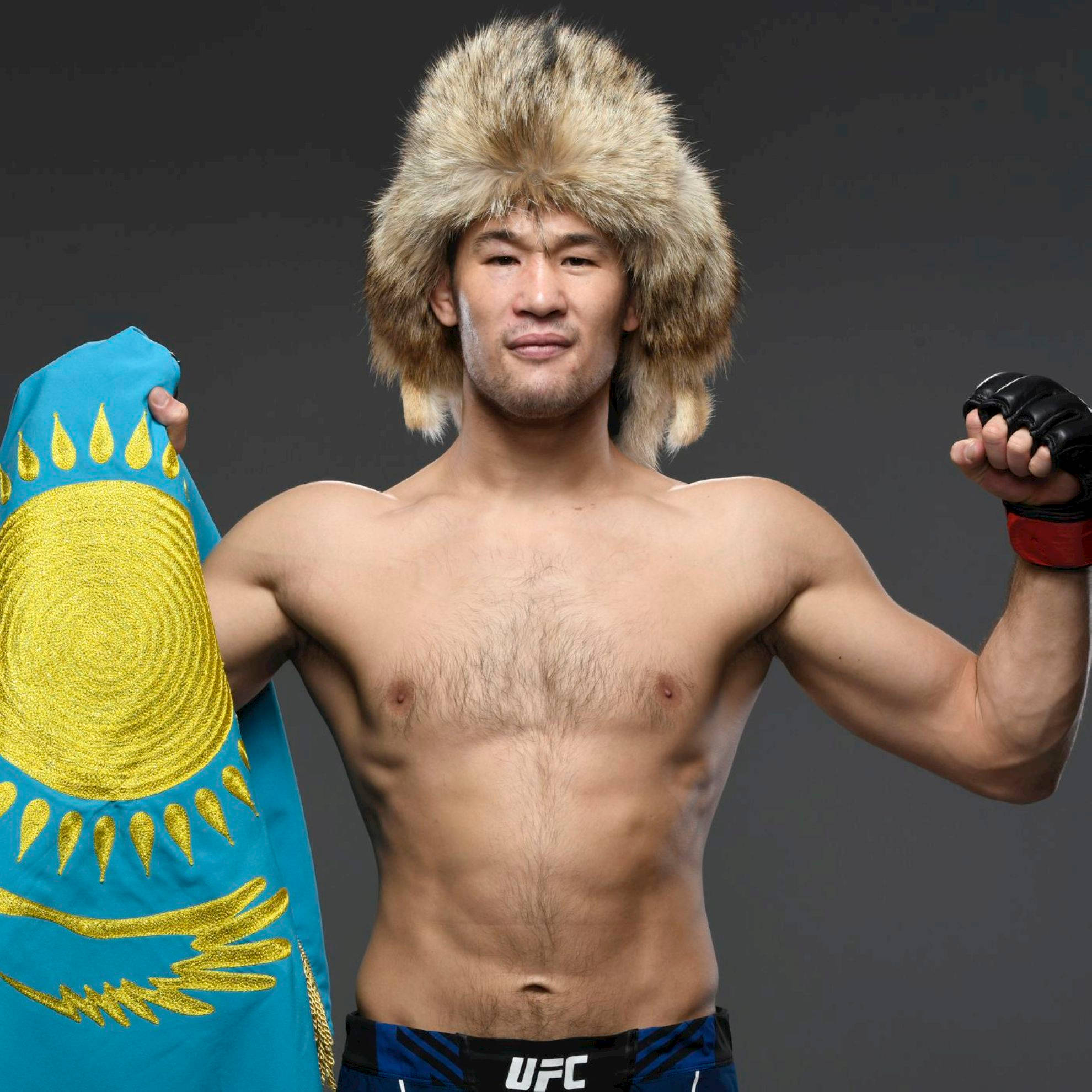 UFC 桌布 壁紙 — 一位肌肉發達的戰士戴著毛帽，自信地在黑暗背景的攝影棚中擺姿勢，手持國旗。