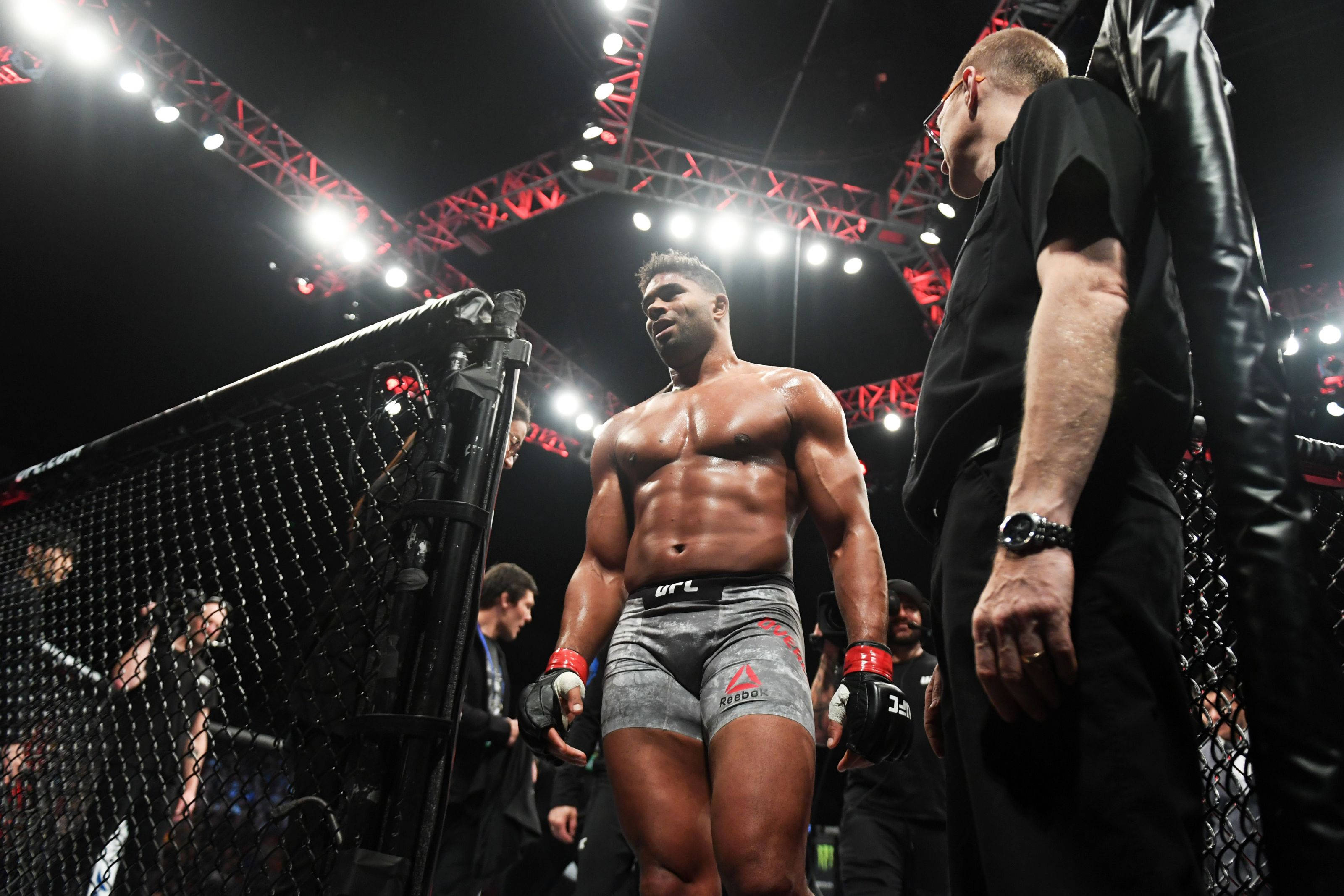 UFC 桌布 壁紙 — 一位肌肉發達的戰士穿著灰色短褲，站在明亮的八角籠內，旁邊有裁判，在明亮的場館燈光下。