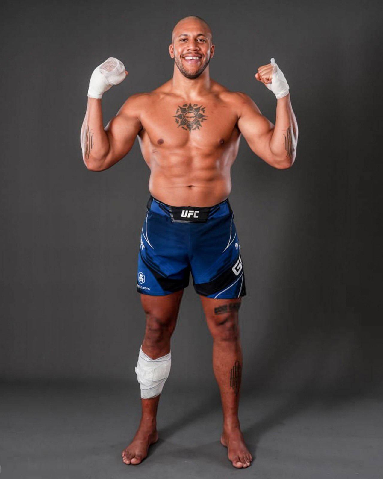 MMA 桌布 壁紙 — 一位肌肉發達的戰士穿著藍色UFC短褲和白色纏手帶，在深灰色工作室背景前自信地舉起拳頭擺姿勢。