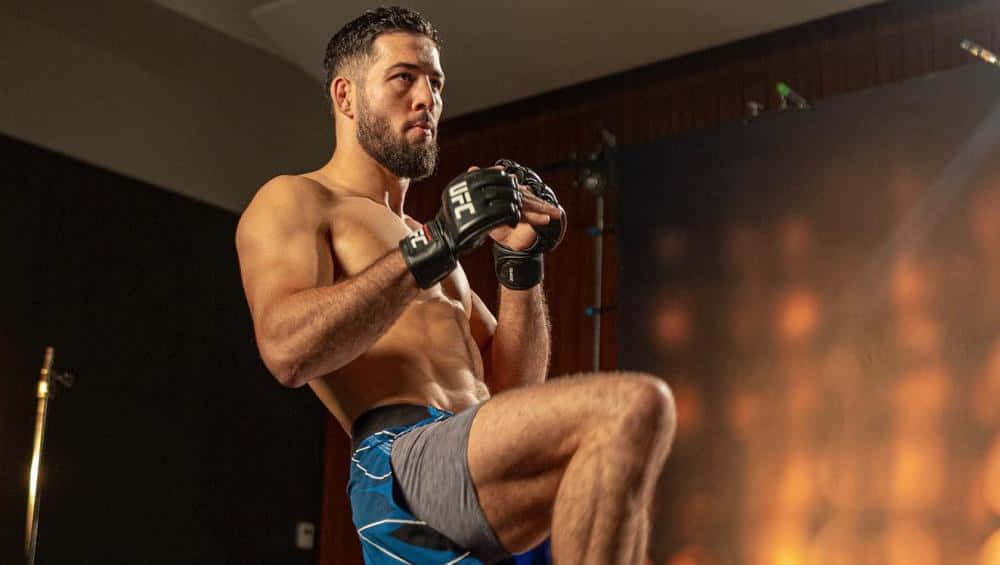 UFC 桌布 壁紙 — 一位肌肉發達的戰士穿著藍色短褲和黑色手套，在溫暖的攝影棚燈光下擺出強有力的姿勢。