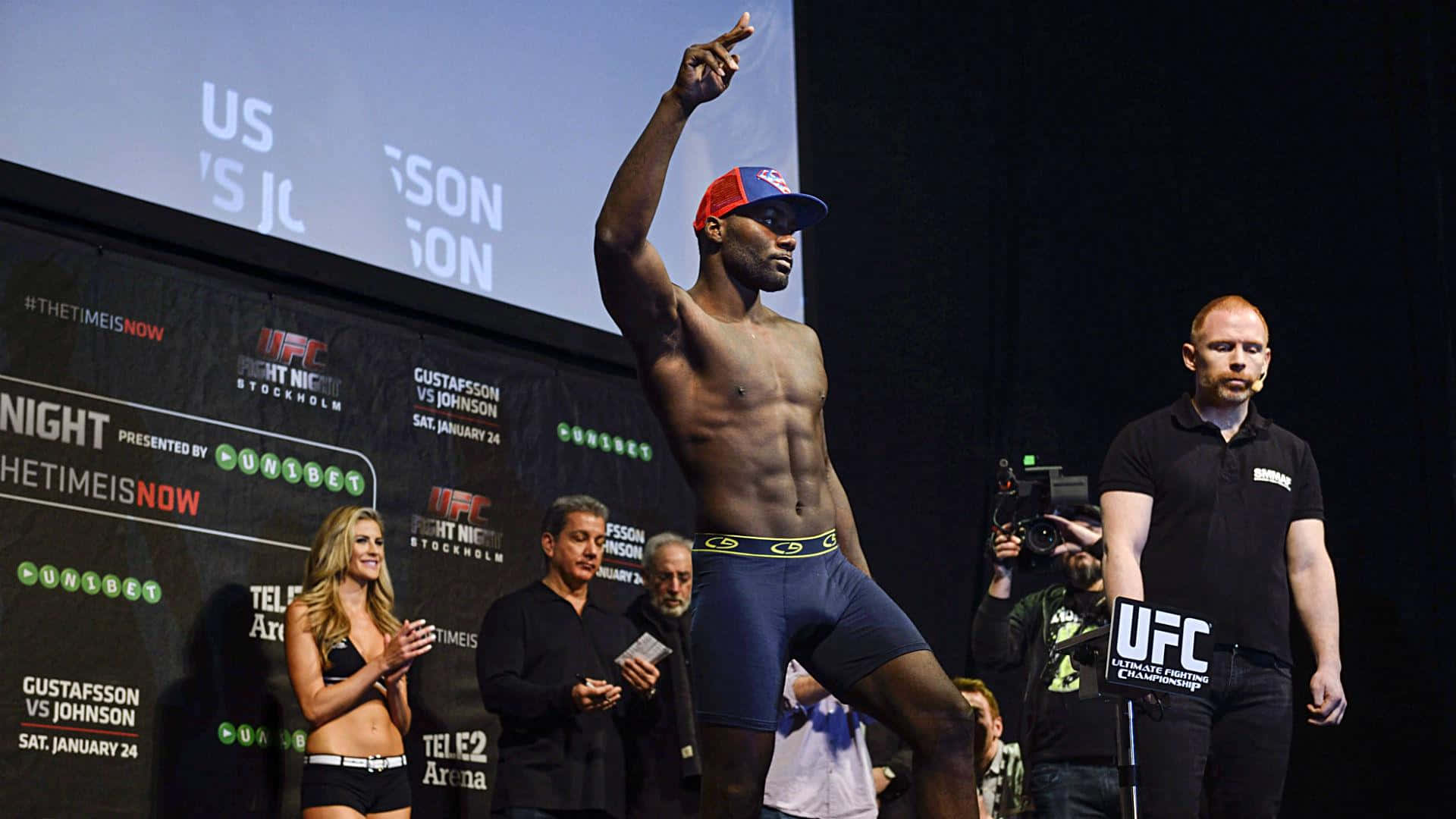 MMA 桌布 壁紙 — 一位肌肉發達的戰士穿著藍色短褲和紅色帽子，在 UFC 稱重舞台上擺出勝利姿勢，背景有觀眾和品牌標誌。