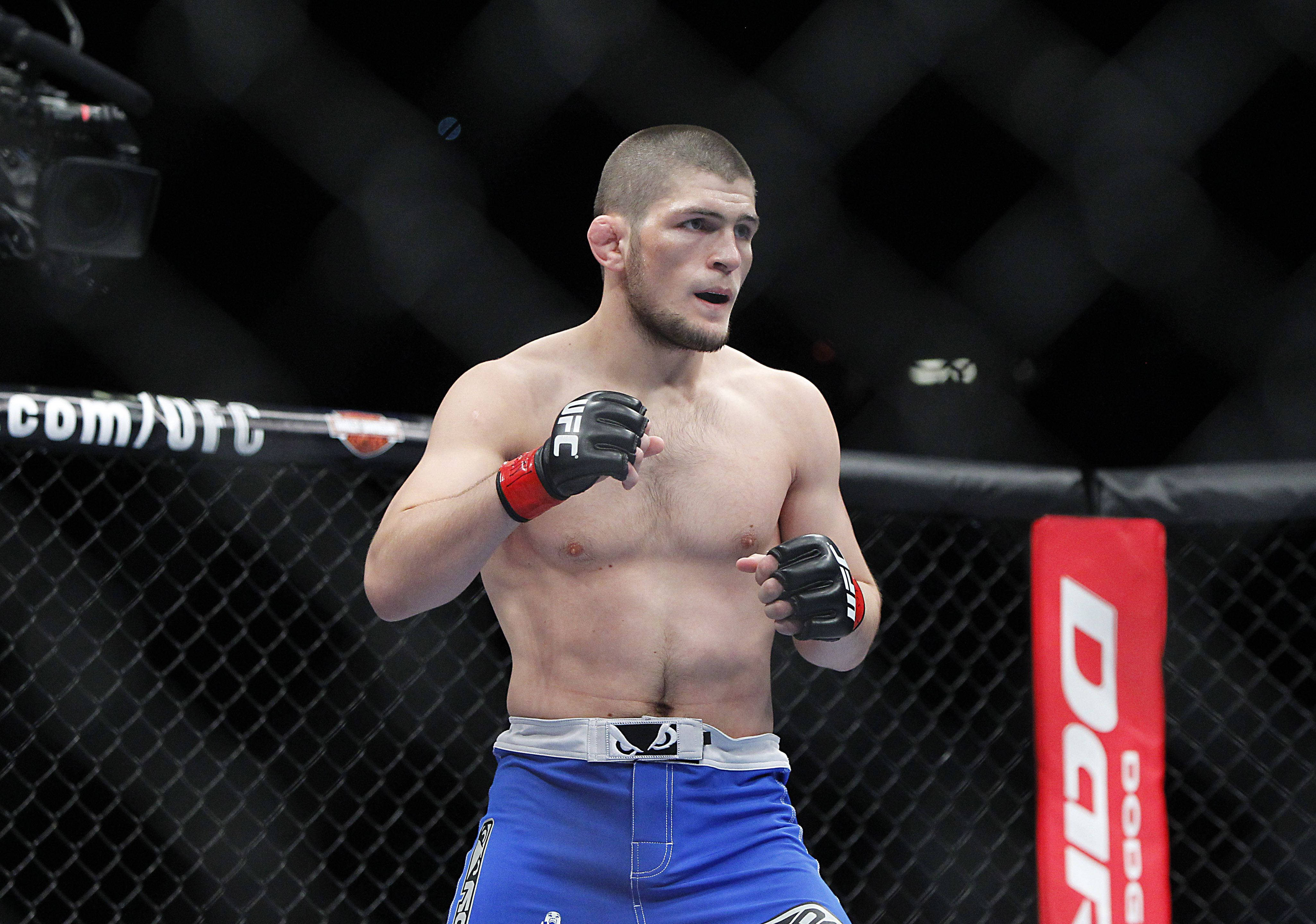 UFC 桌布 壁紙 — 一位肌肉發達的戰士穿著藍色短褲和黑色手套，在UFC八角籠內警覺地站立，鐵絲網圍欄和攝影機清晰可見。