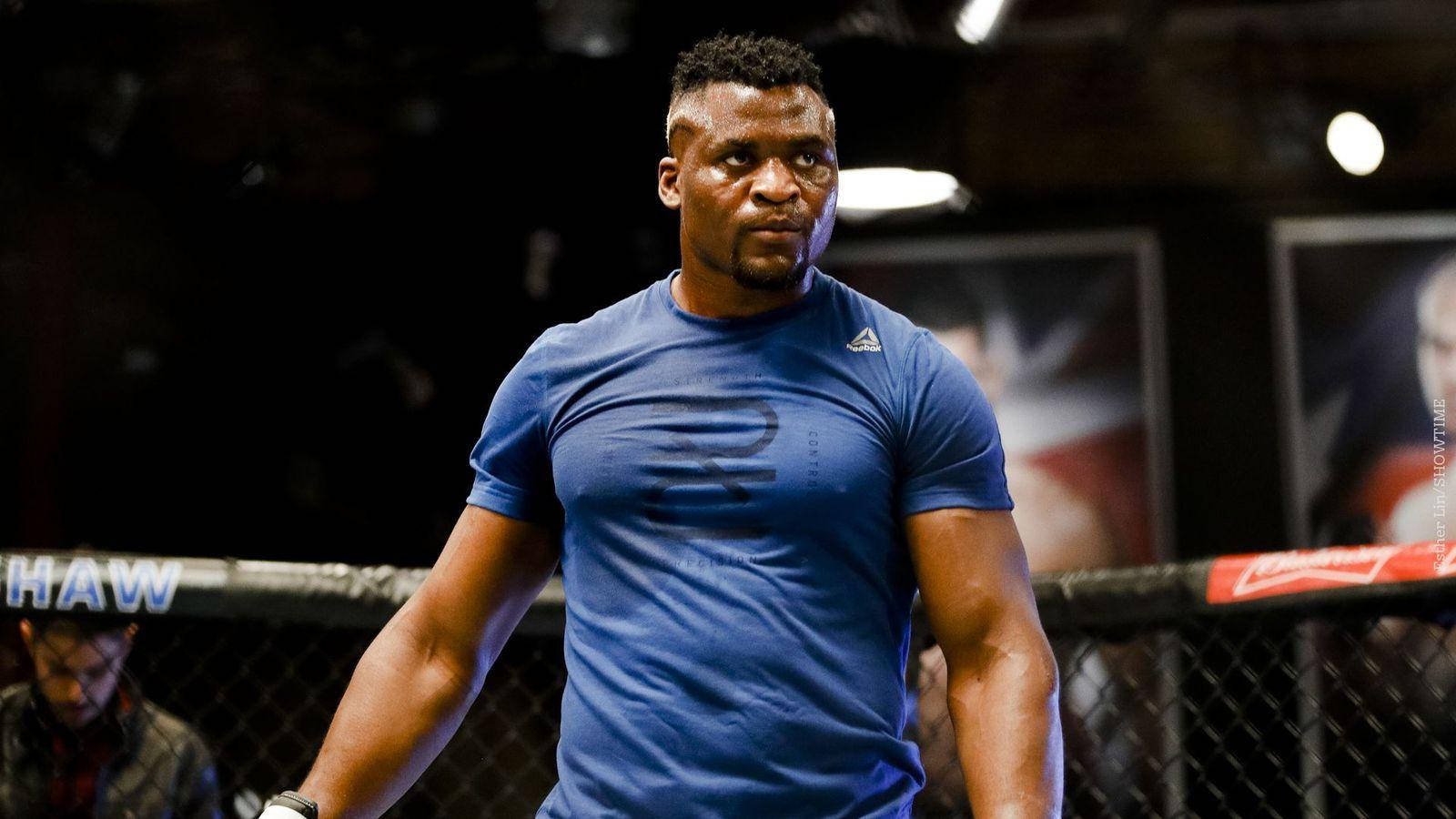 MMA 桌布 壁紙 — 一位肌肉發達的戰士穿著藍色襯衫，在光線昏暗的MMA八角籠內，帶有鐵絲網圍欄和贊助商標誌，神情專注。