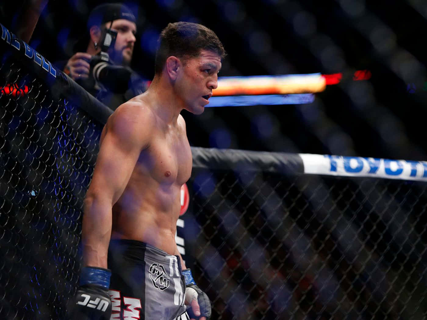 UFC 桌布 壁紙 — 一位肌肉發達的戰士站在UFC八角籠內，在明亮的場館燈光下展現專注的強烈氣勢。