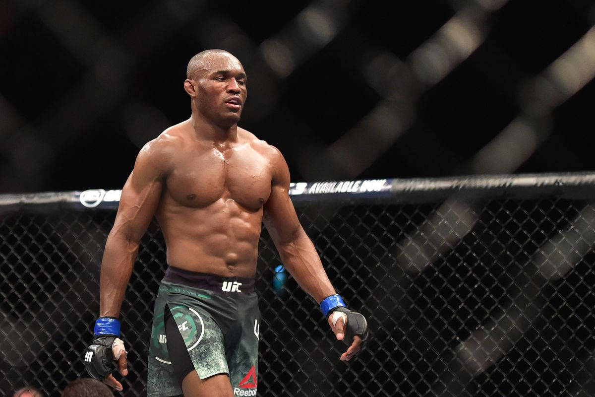 UFC 桌布 壁紙 — 一位肌肉發達的戰士站在燈光昏暗的八角籠內，汗水在他結實的軀幹和專注的表情上閃閃發光。