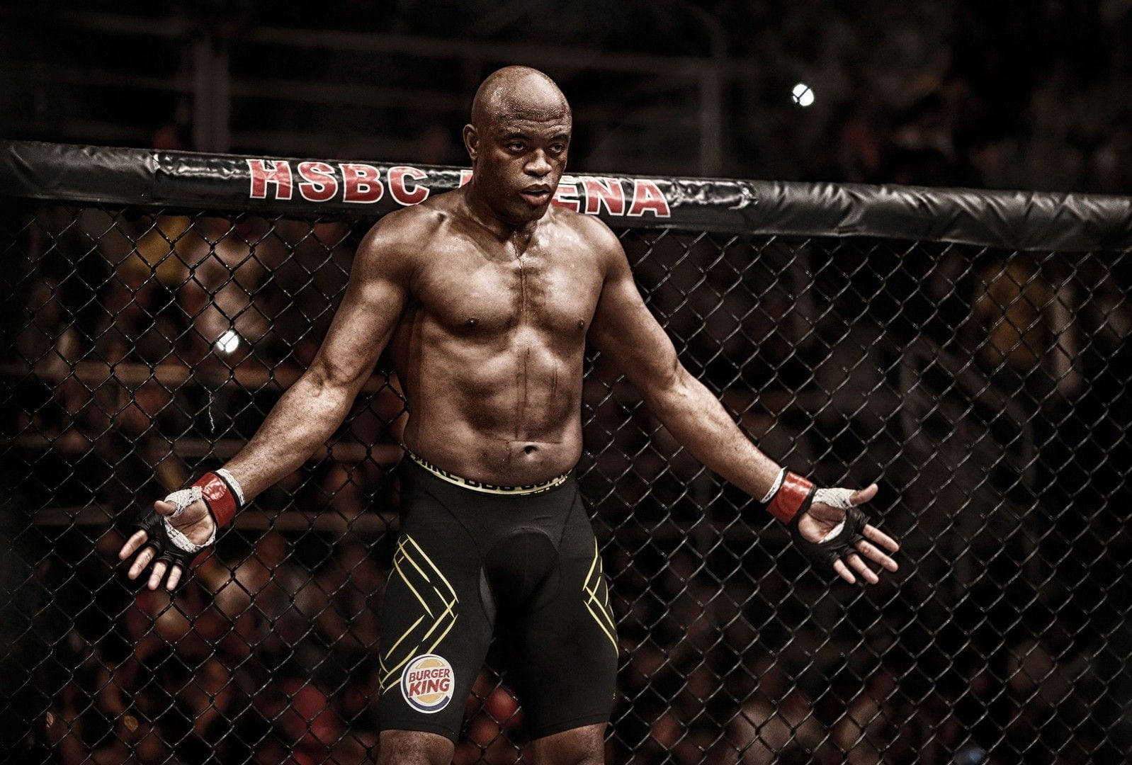 UFC 桌布 壁紙 — 一位肌肉發達的戰士站在籠內，雙臂伸展，在明亮的競技場燈光下。