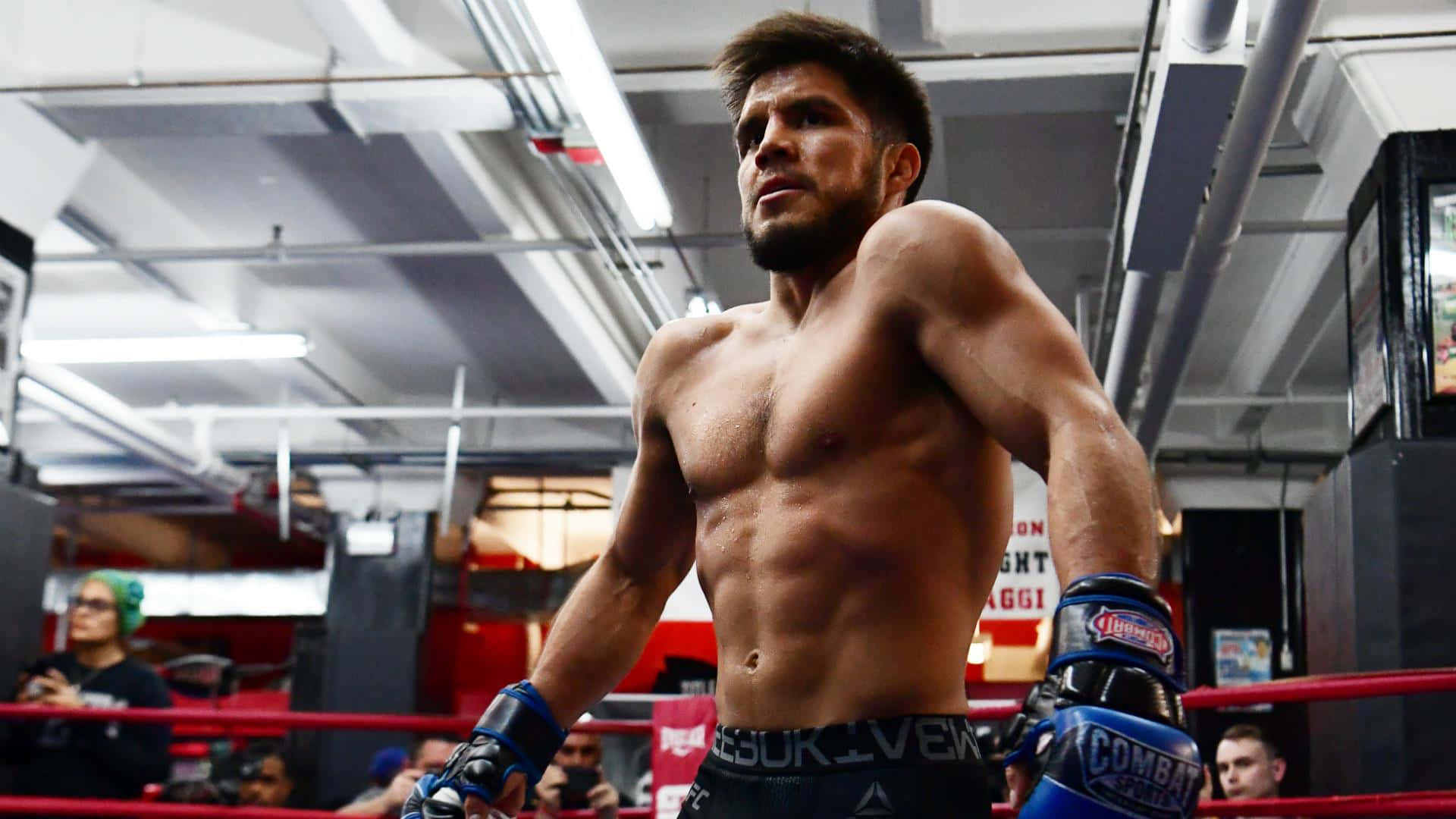 UFC 桌布 壁紙 — 一位肌肉發達的戰士赤裸上身站在拳擊台上，紅色繩索和藍色手套在明亮的頭頂燈光下。