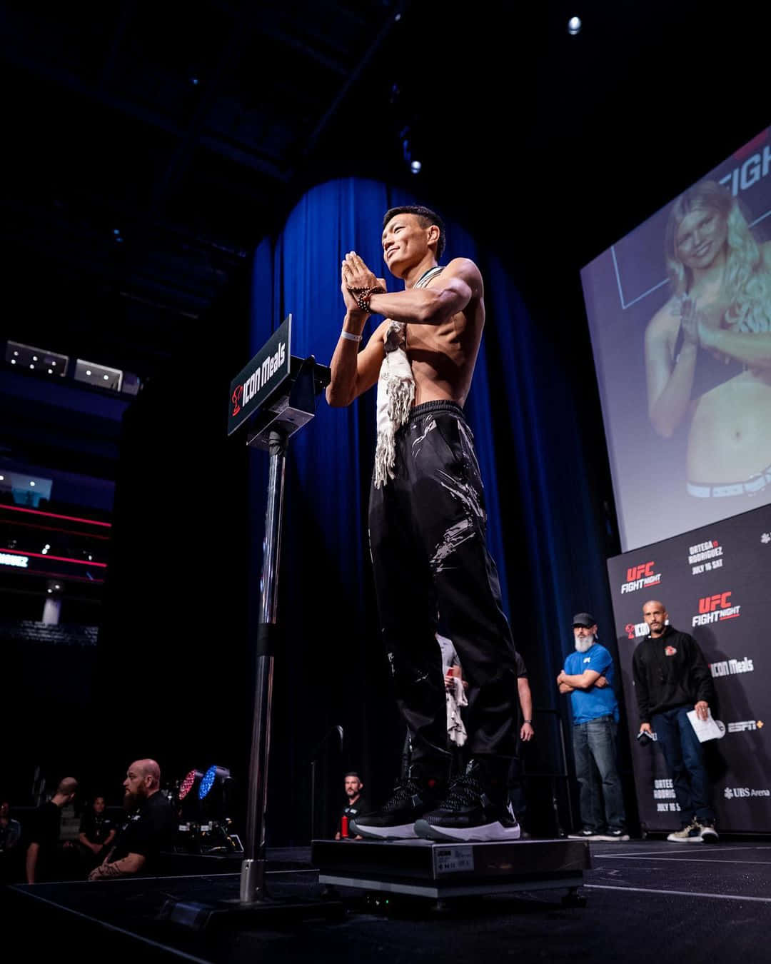 MMA 桌布 壁紙 — 一位肌肉發達的戰士赤裸上身站在體重秤上，在明亮的舞台燈光下進行UFC稱重，身後有一個大螢幕。