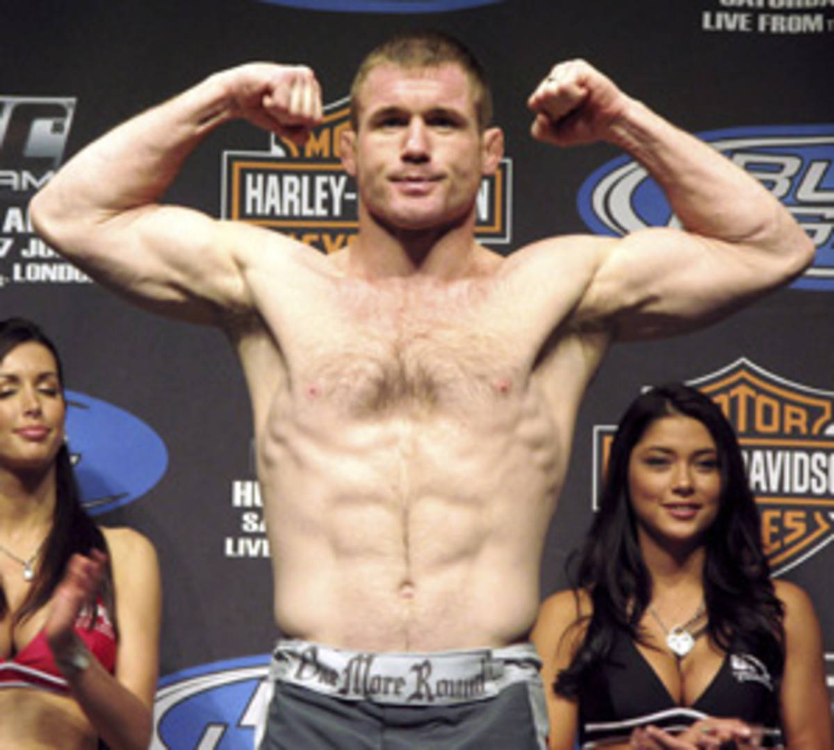 UFC 桌布 壁紙 — 一位肌肉發達的拳手在UFC稱重活動中在磅秤上彎曲手臂，旁邊有兩位身穿比基尼的女性。