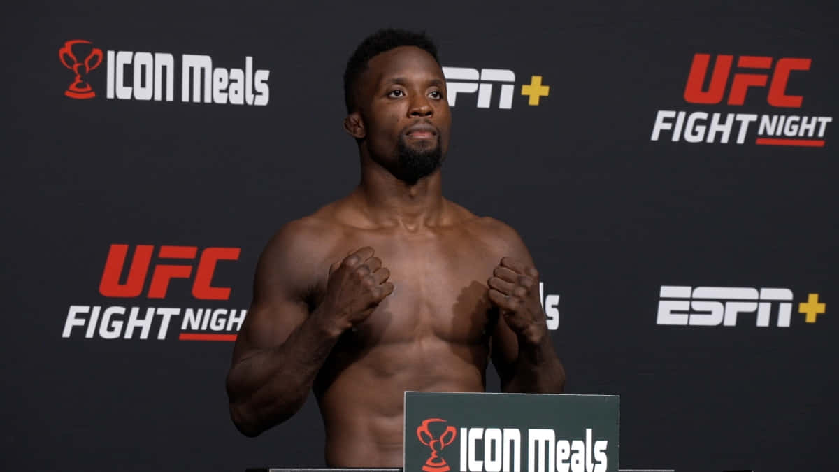 UFC 桌布 壁紙 — 一位肌肉發達的拳手在UFC記者會上赤裸上身，舉起拳頭，背景帶有品牌標誌。