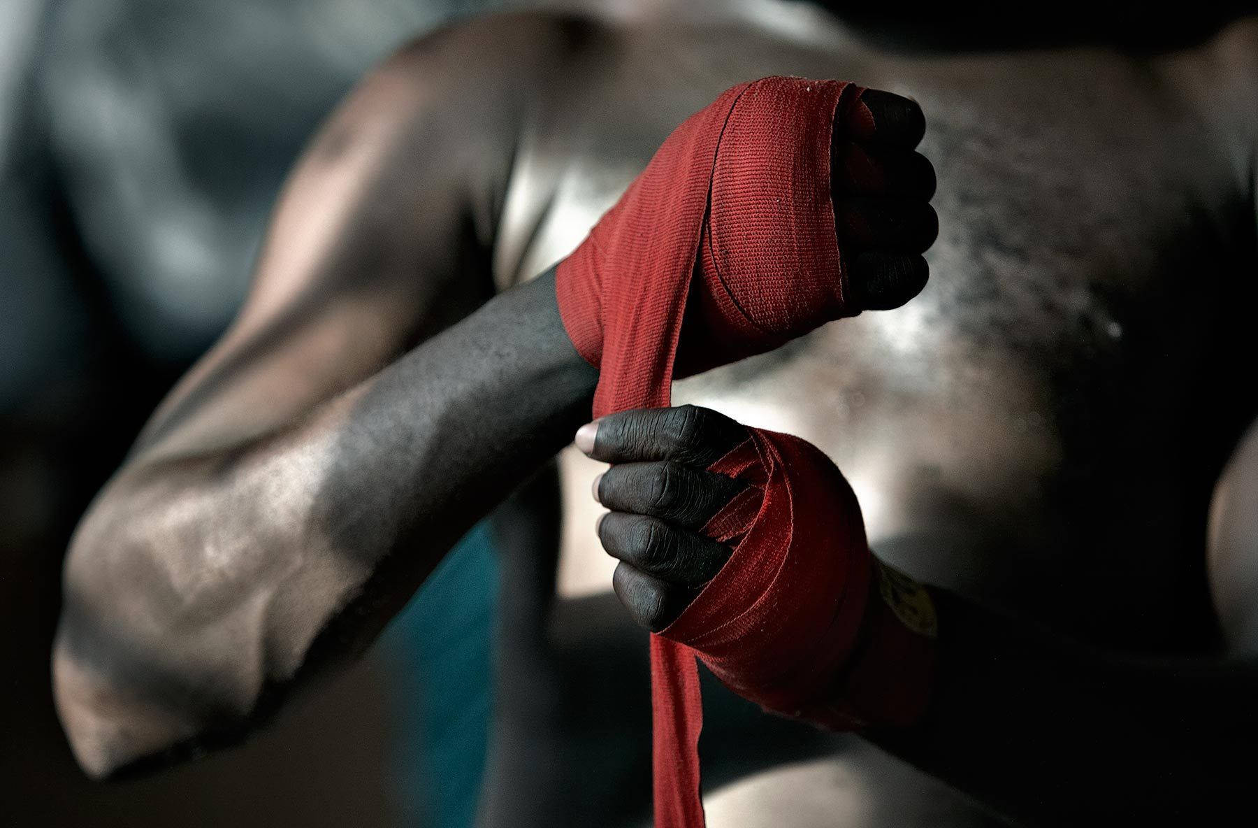 Boxing 桌布 壁紙 — 一位肌肉發達的拳手在燈光昏暗的健身房中，用戲劇性的燈光將紅色纏手帶纏繞在拳頭上。